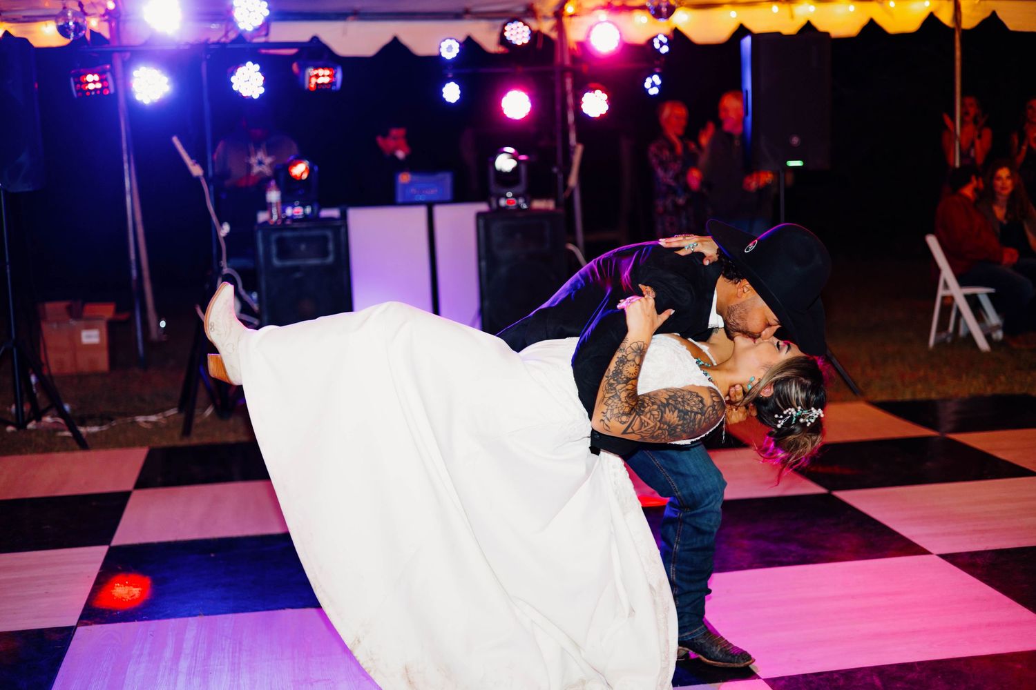 Groom lifts bride in white gown during first dance on checkered floor at outdoor wedding reception.