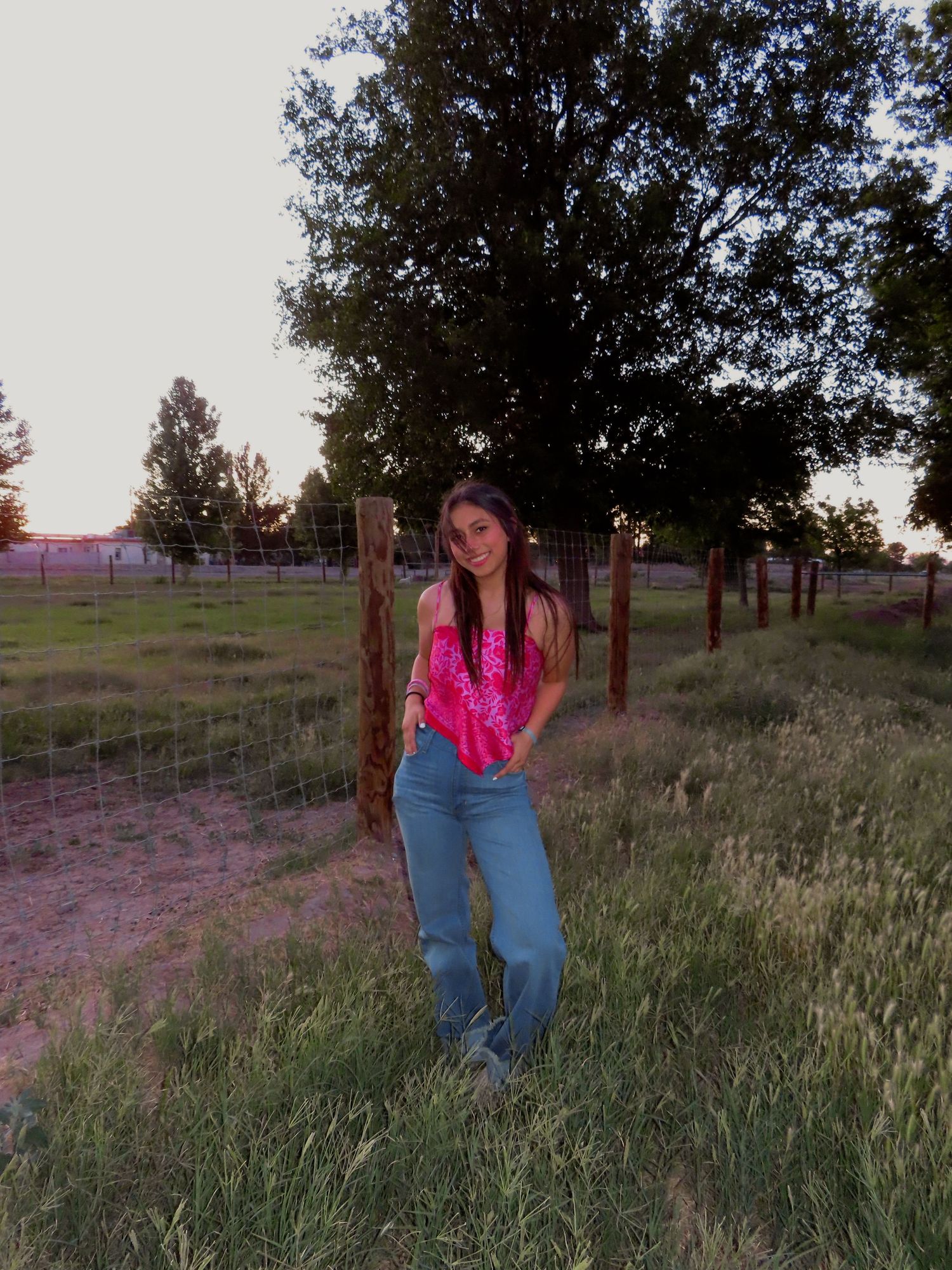 Young woman in pink top and jeans posing near a fence in a rural field at sunset.