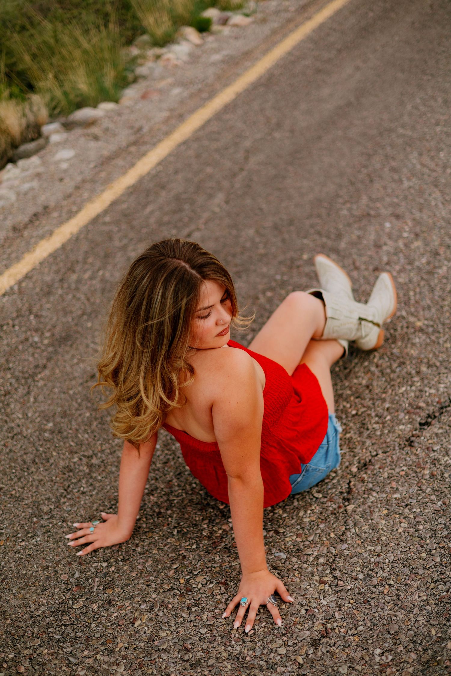 Woman in red top, denim shorts, and white cowboy boots posing on a road at sunset.