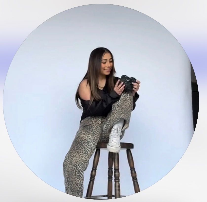 Woman in black top and leopard print pants sits on stool holding camera in a circular framed studio photo.