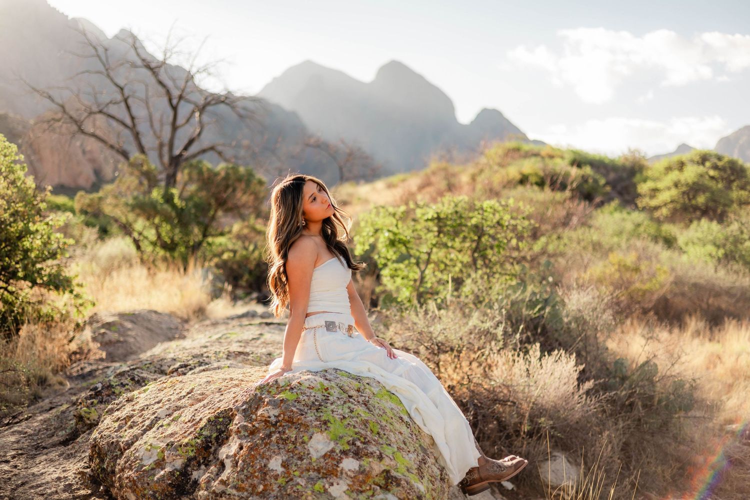 Woman in white dress sitting on boulder surrounded by lush mountain landscape in golden sunlight.