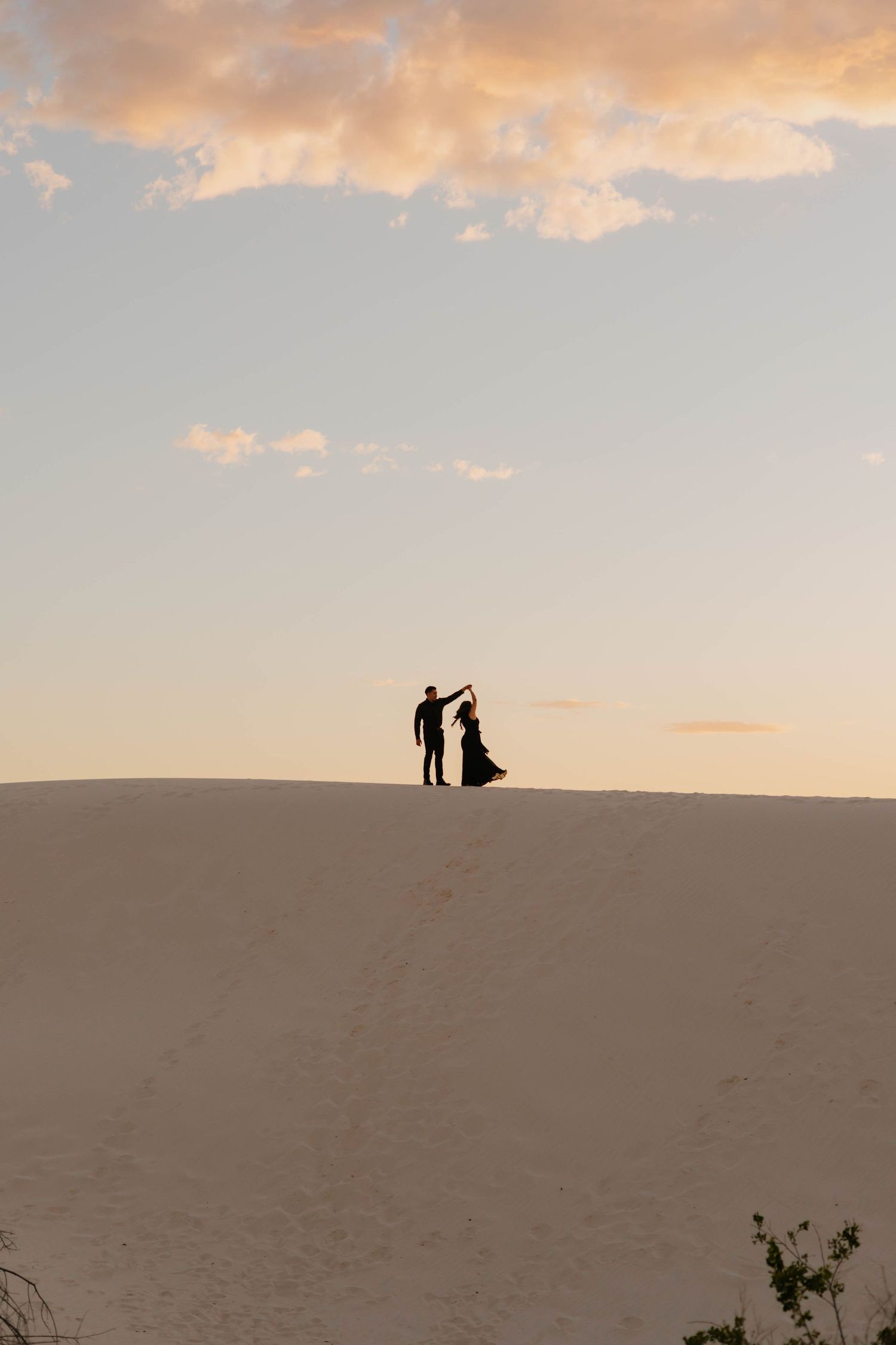 Silhouette of couple dancing on white sand dunes at sunset with dramatic cloudy sky backdrop.