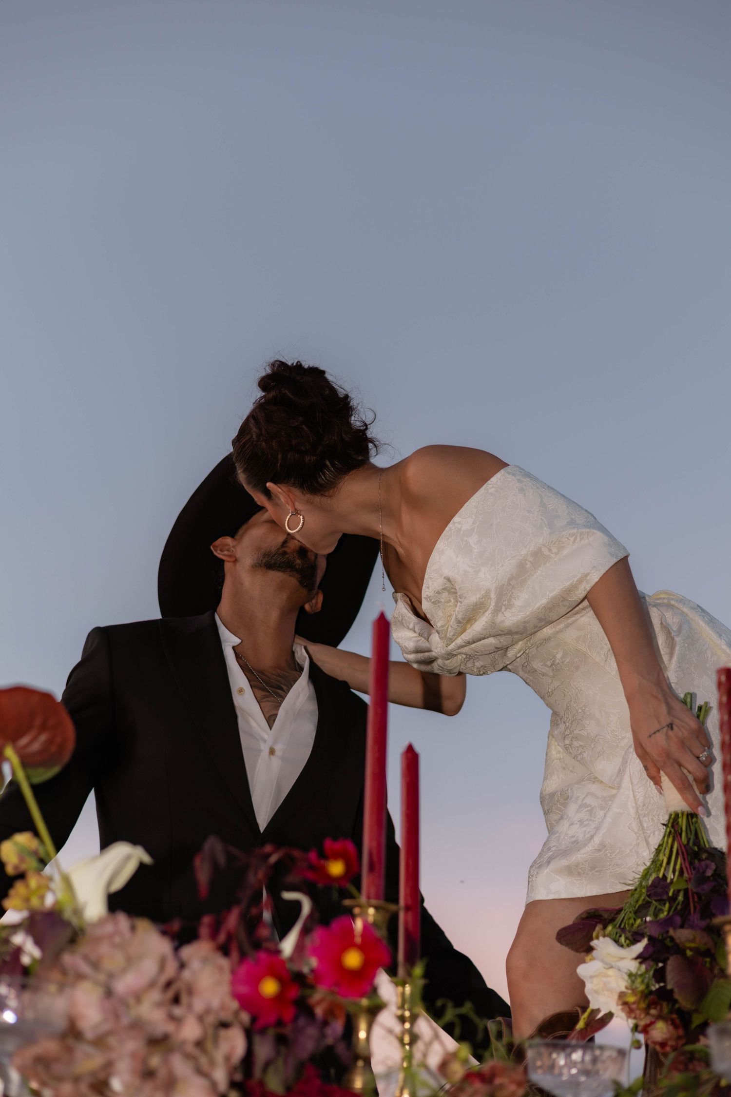 Bride in white dress leans down to kiss groom in black suit at outdoor wedding reception with red candles and floral arrangements.
