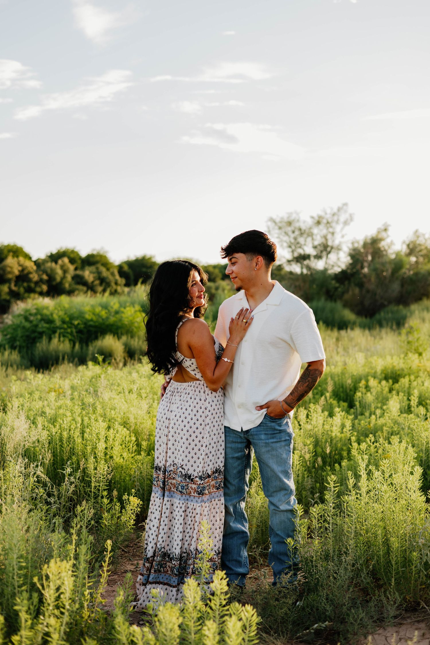Couple embracing in a lush green field at golden hour, woman in floral maxi dress, man in white shirt and jeans.