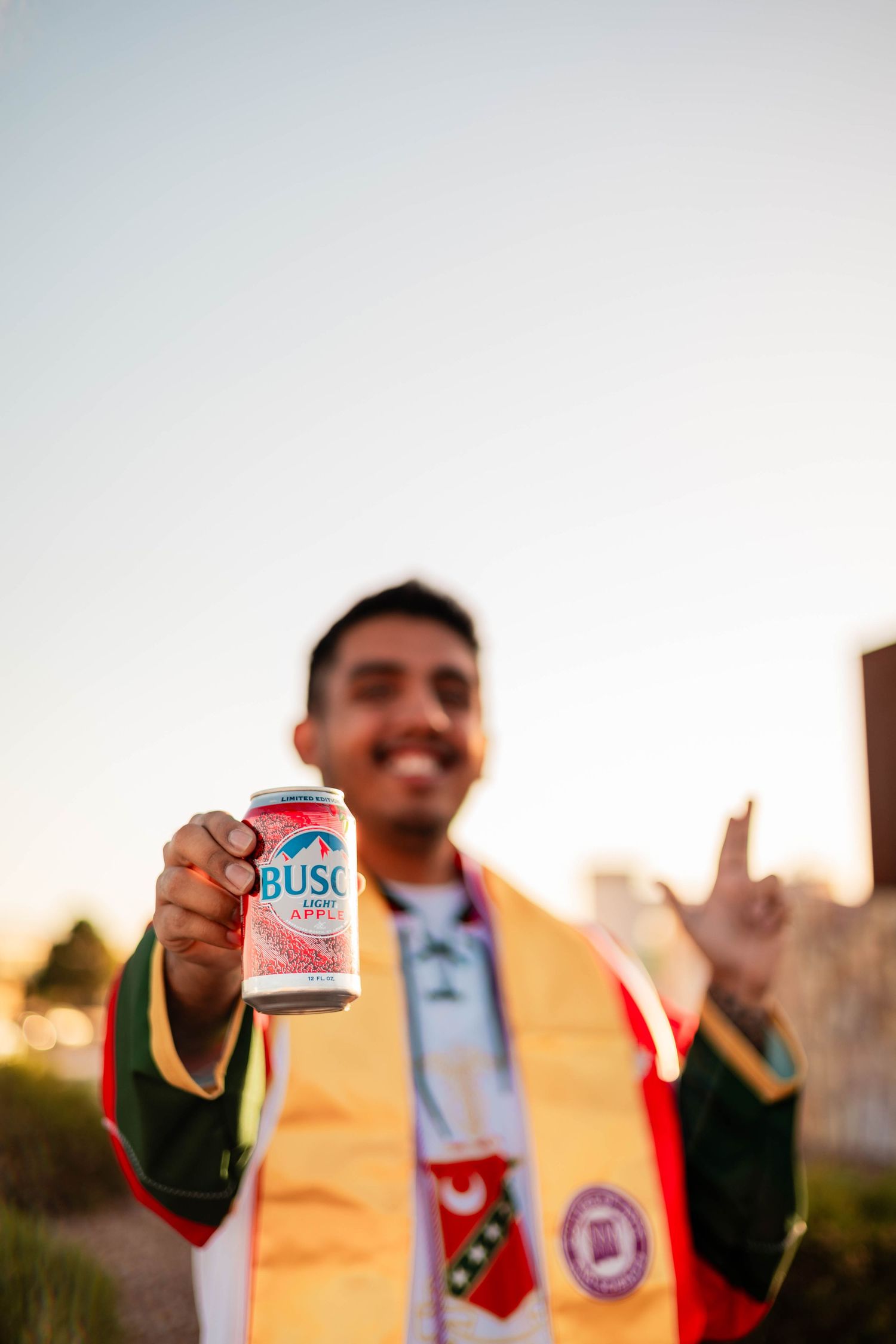 Smiling graduate in colorful sash holds Busch beer can toward camera at golden hour.