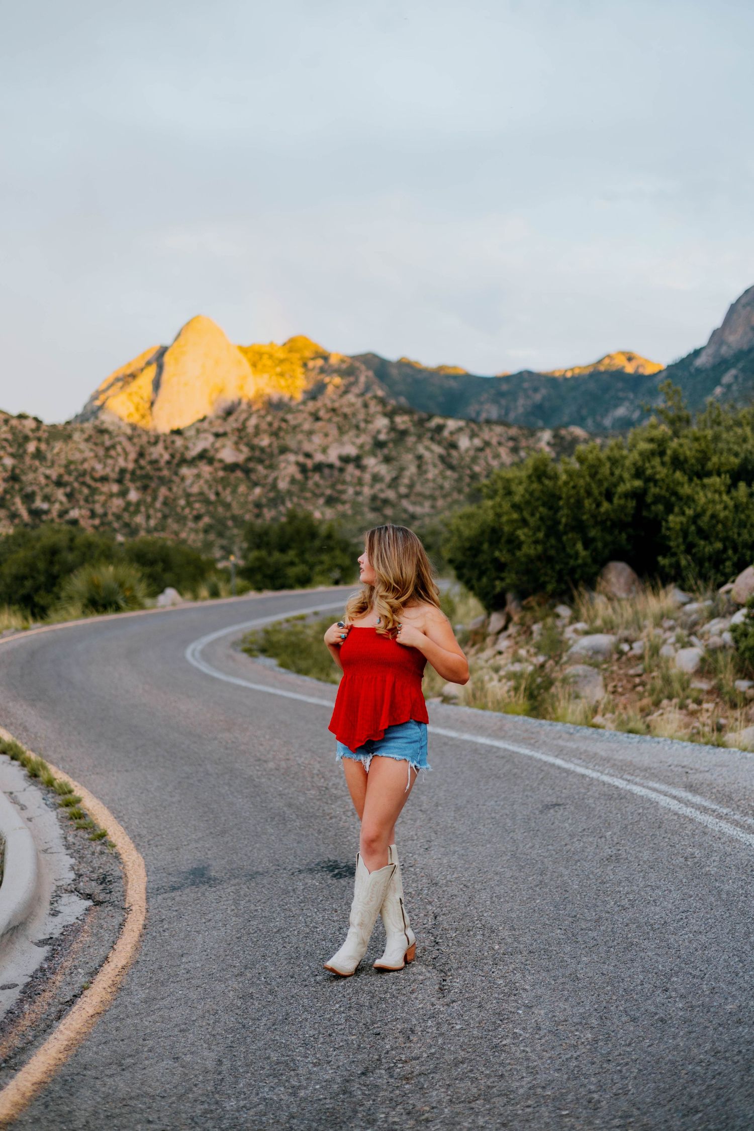 Woman in red top, denim shorts, and white cowboy boots standing on a winding road with mountain scenery at sunset.