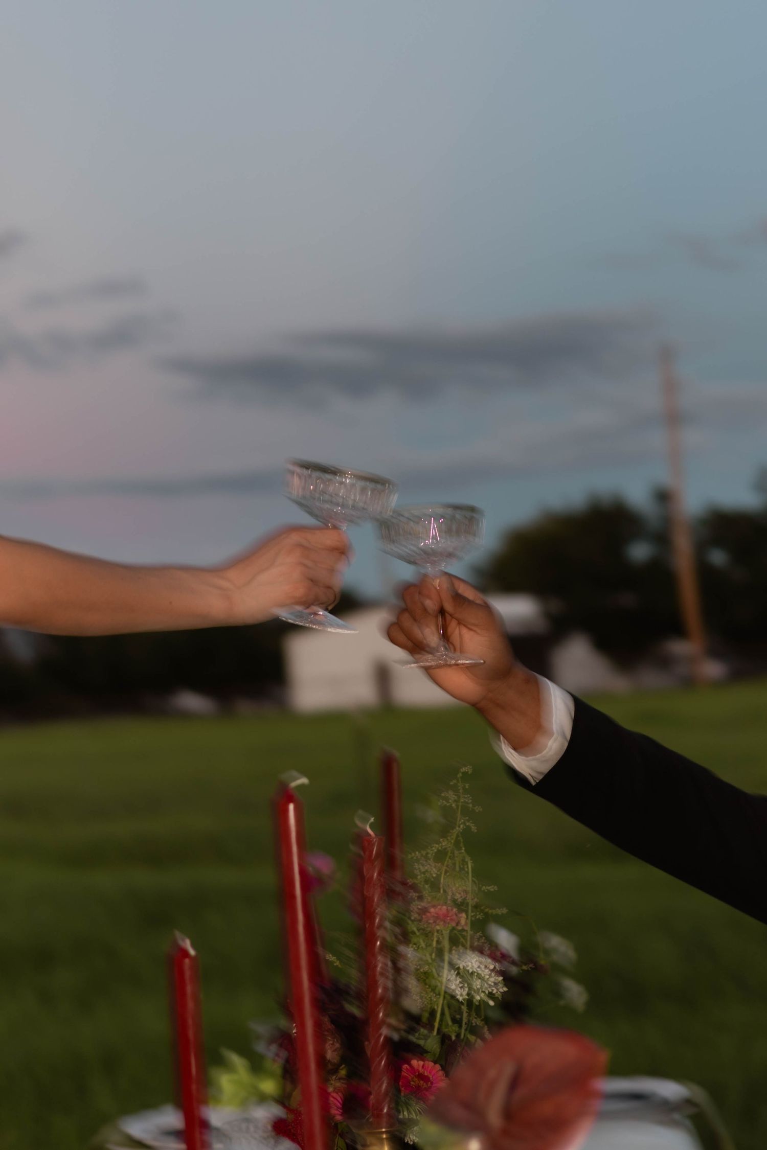 Two hands clinking crystal champagne coupes at an outdoor wedding reception at dusk with red candles and floral centerpiece.