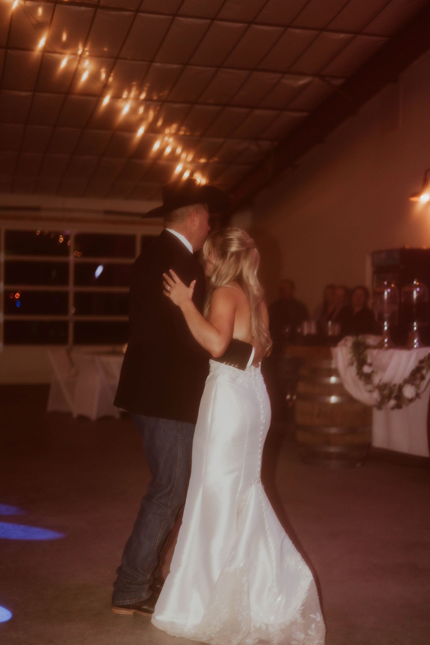 Bride and groom share first dance in dimly lit wedding venue with warm string lights overhead.