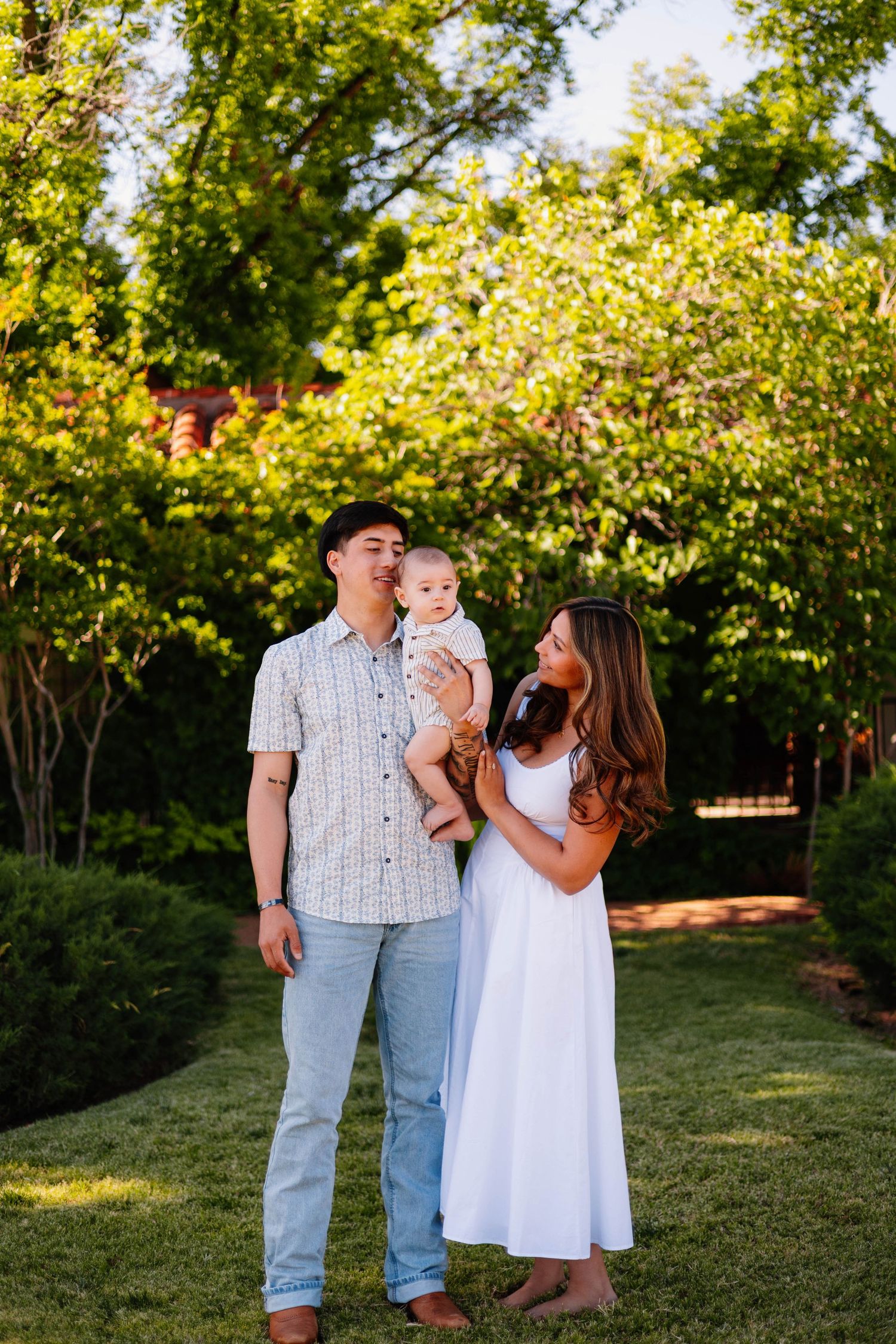 Young couple holding baby in lush green garden during warm golden hour sunlight session.