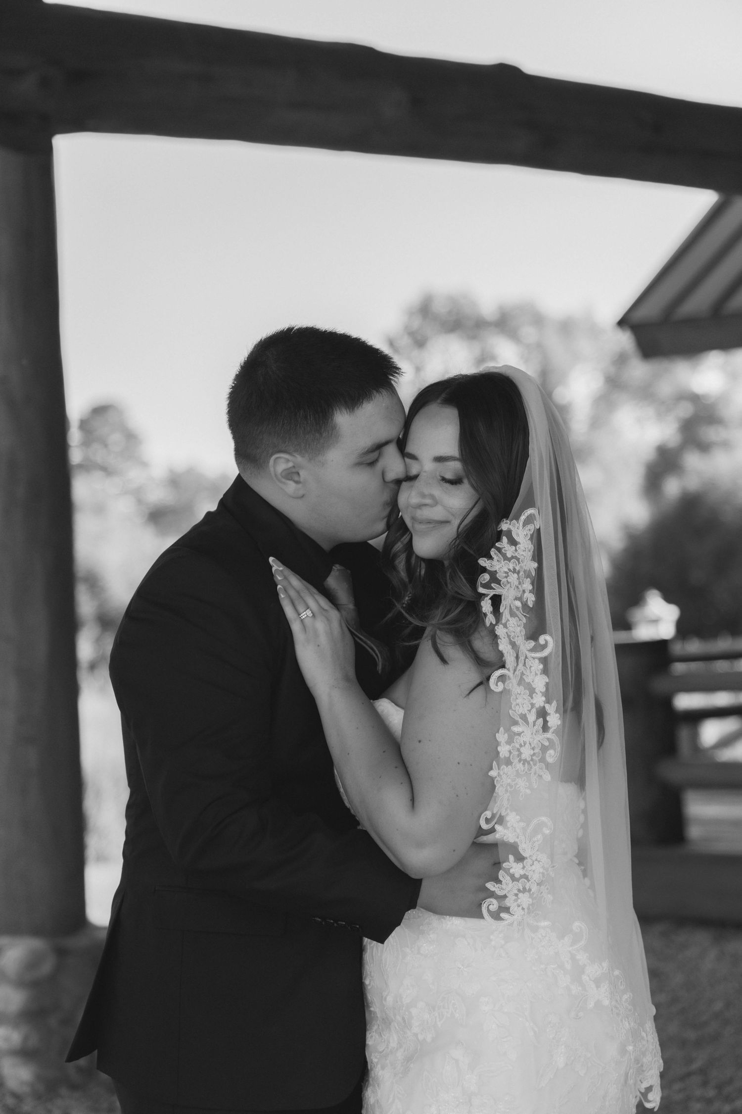 Black and white wedding photo of groom kissing bride on cheek, bride wearing lace veil, under wooden pergola.