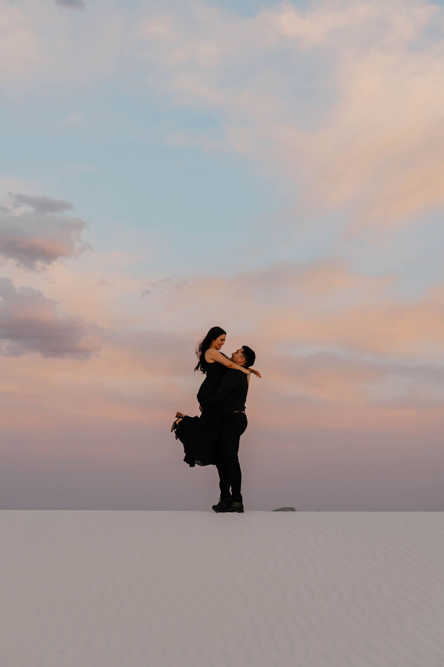 Romantic couple embracing on white sand dunes beneath a pastel sunset sky.