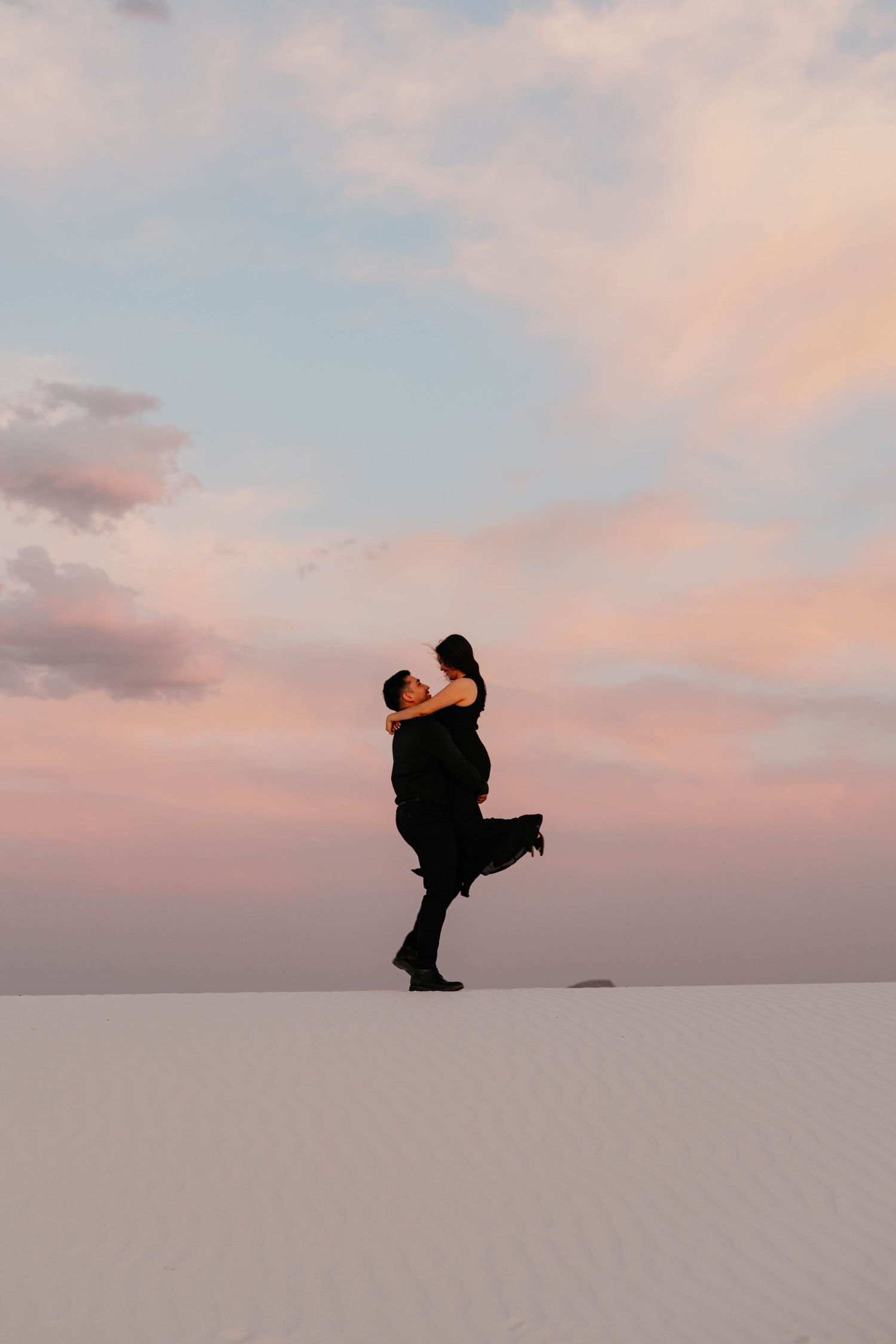 Couple sharing a romantic moment at sunset on white sand dunes, man lifting woman against a pastel sky.