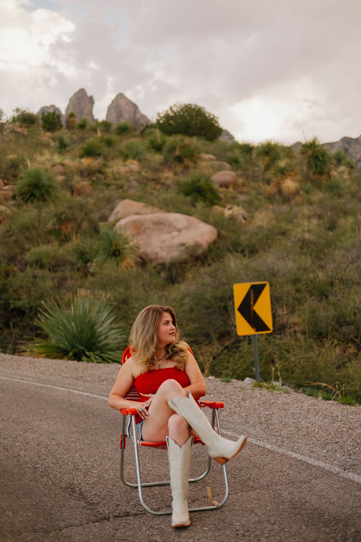Woman in red dress and white cowboy boots sits on a folding chair on a desert road with rocky hills behind her.