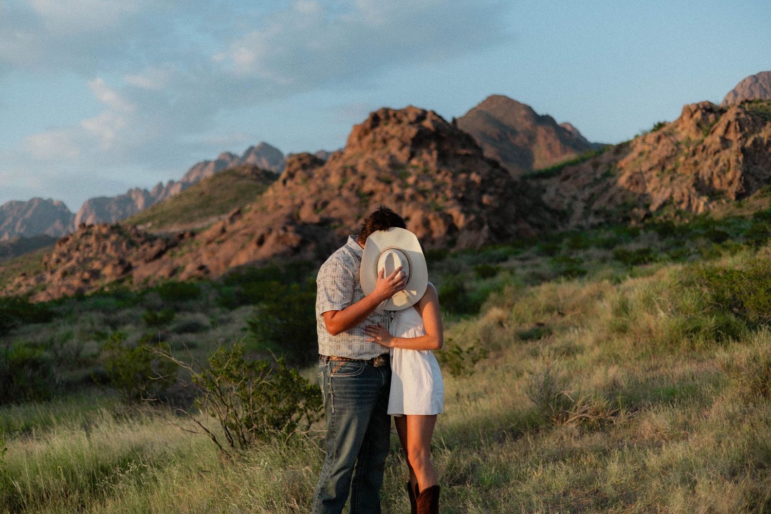 Couple embracing in a lush desert landscape with rugged red rock mountains in the background at golden hour.