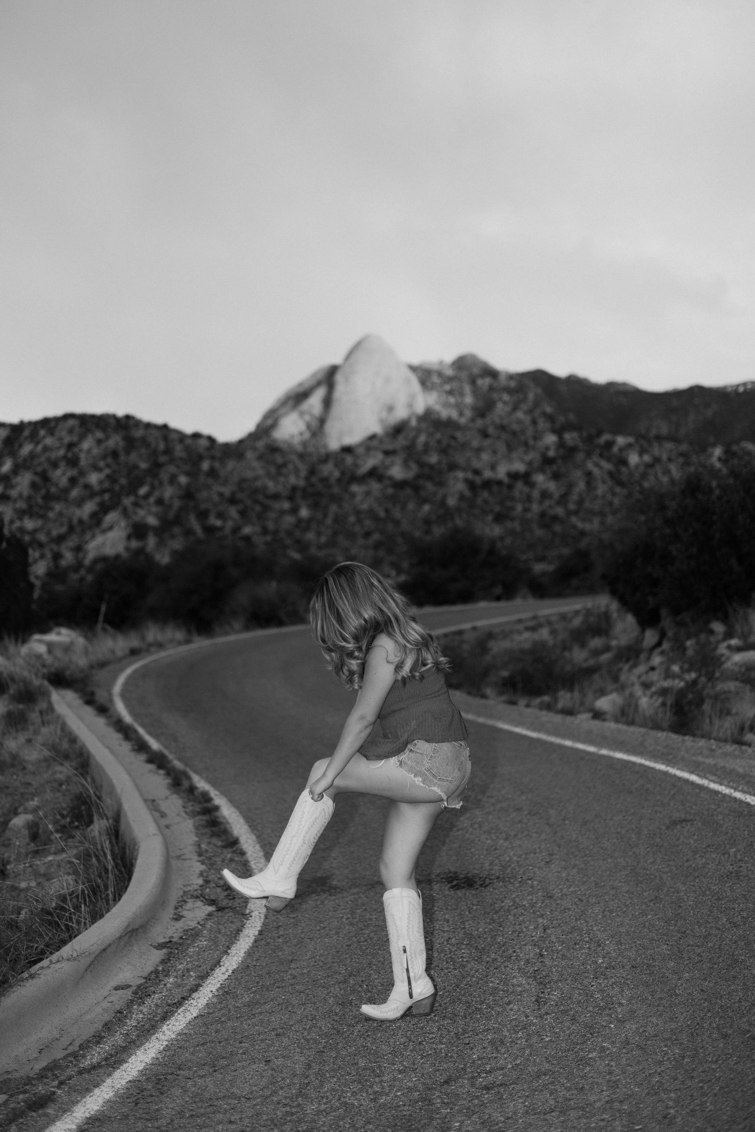 Black and white photo of woman adjusting white cowboy boots on a winding mountain road.