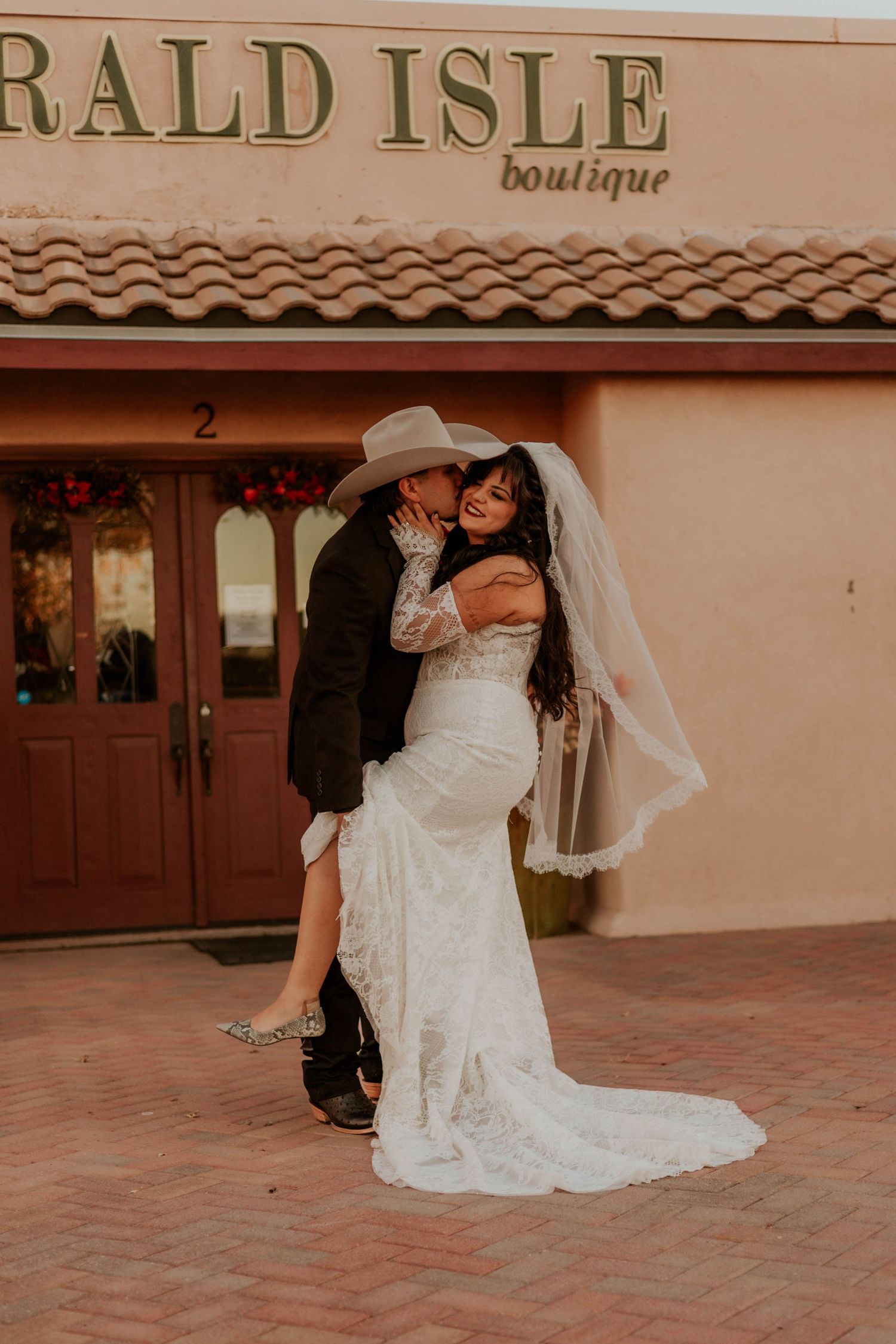Bride in lace gown and veil embraces groom in cowboy hat outside Emerald Isle Boutique on their wedding day.