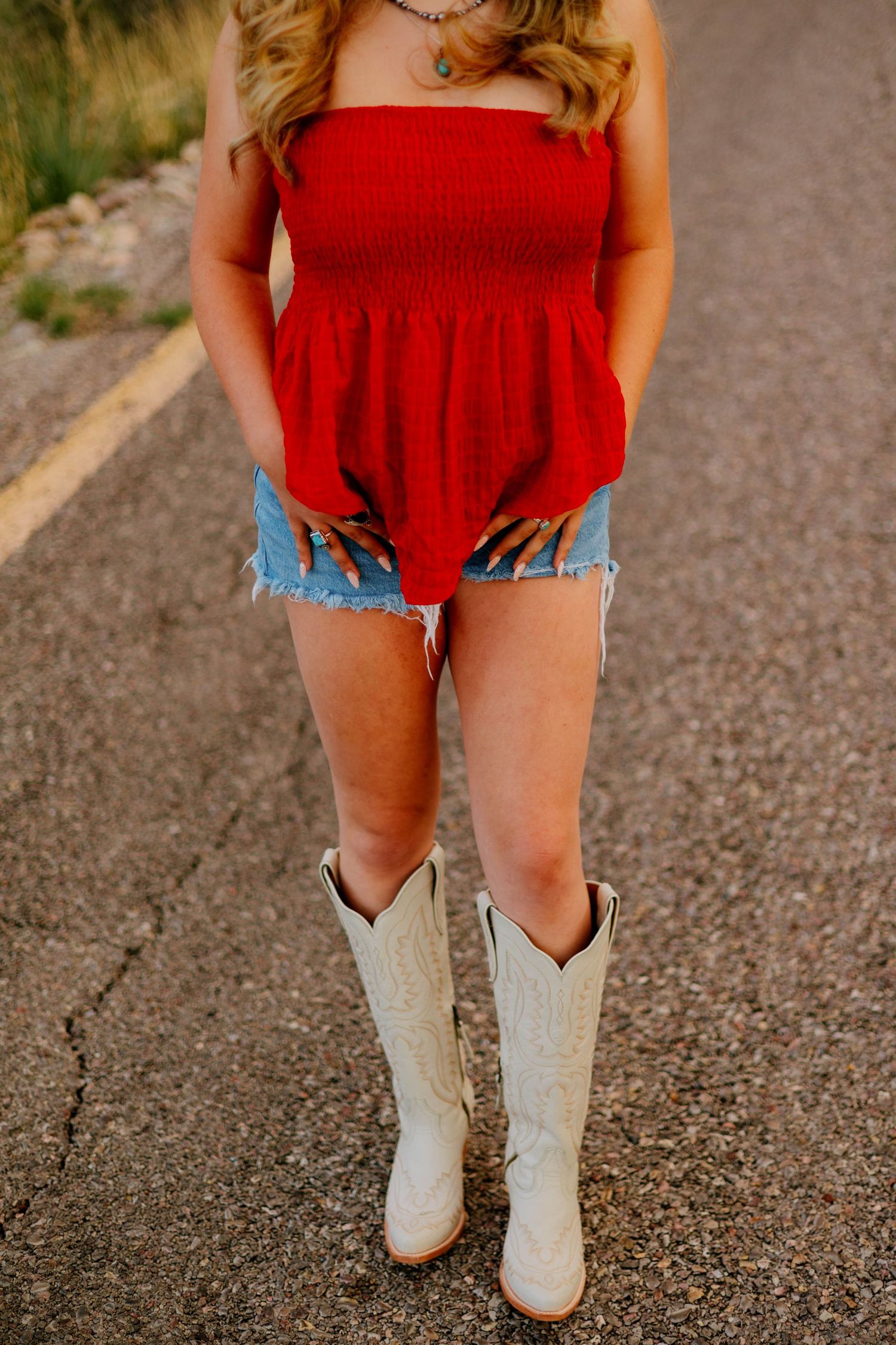 Woman in red smocked tube top, denim cutoff shorts, and white cowboy boots standing on a road.