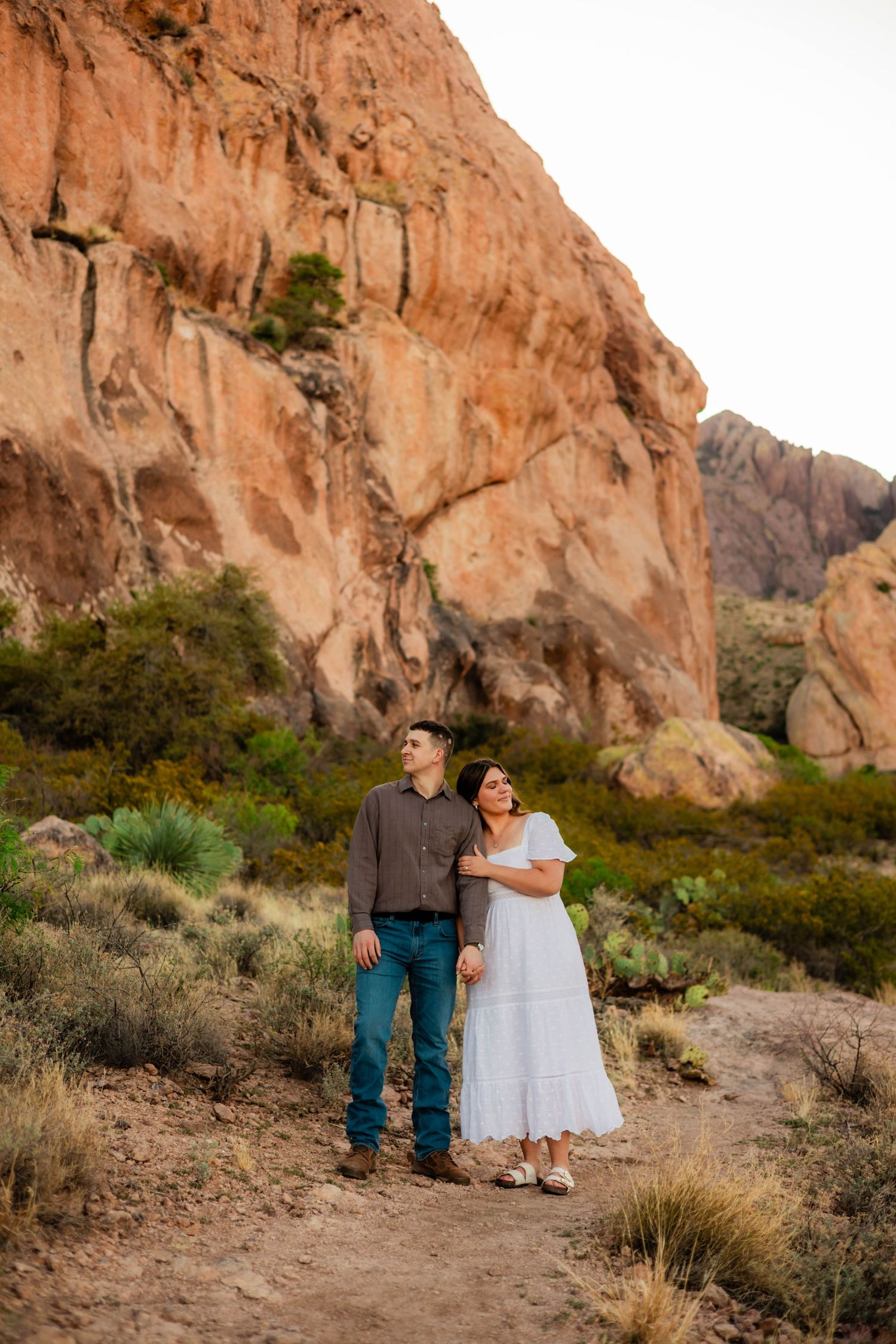 Couple standing together on desert trail gazing up at dramatic red rock canyon walls at dusk.
