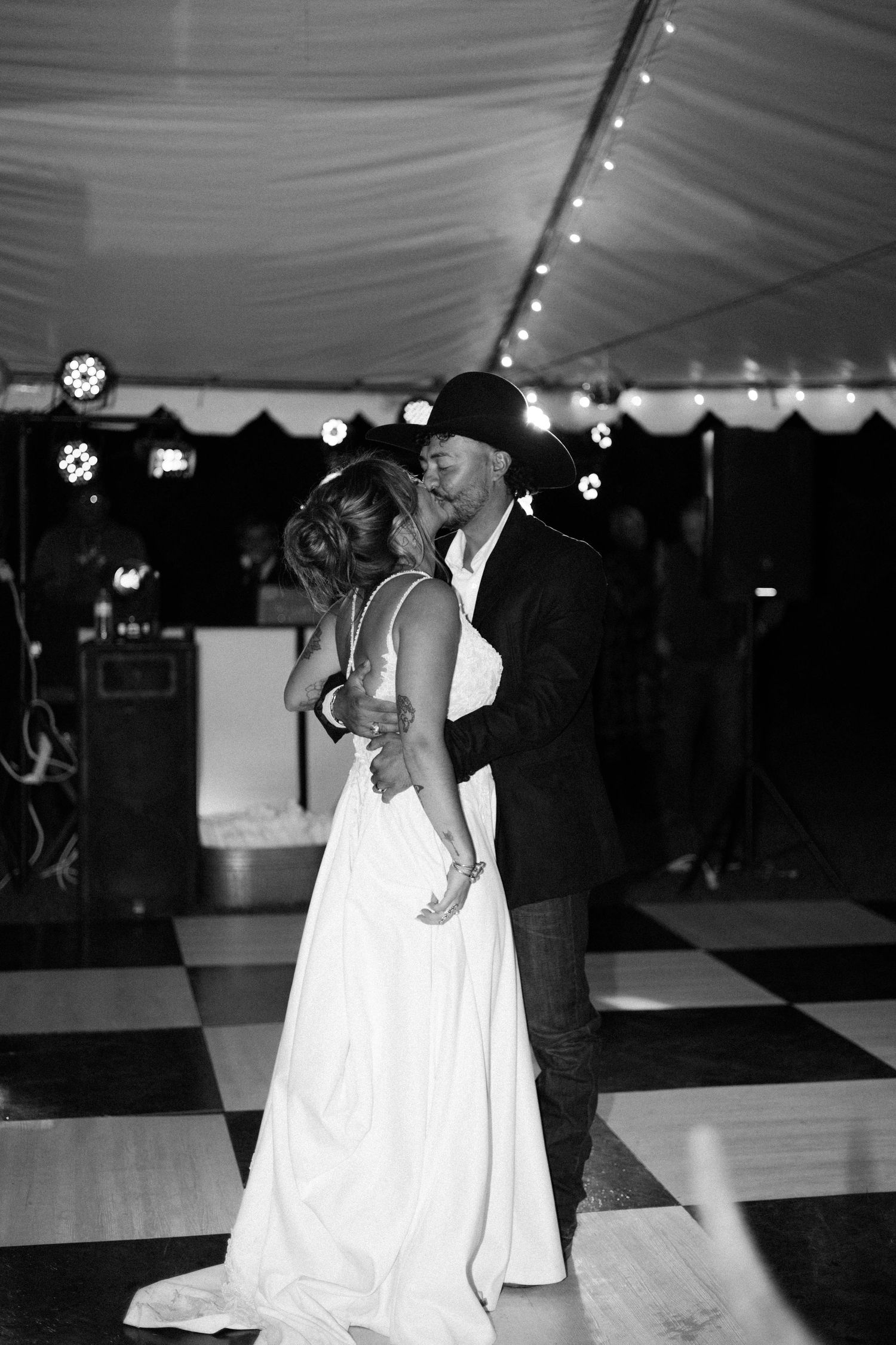 Black and white photo of a bride and groom sharing their first dance kiss under twinkling lights at wedding reception.