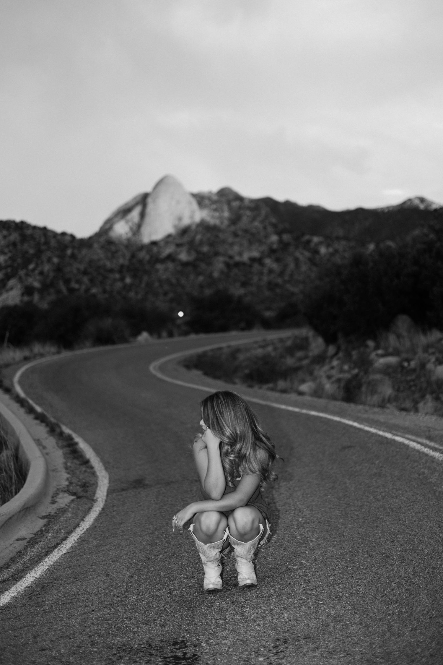 Black and white photo of a young woman crouching on a winding road with mountains in the background.