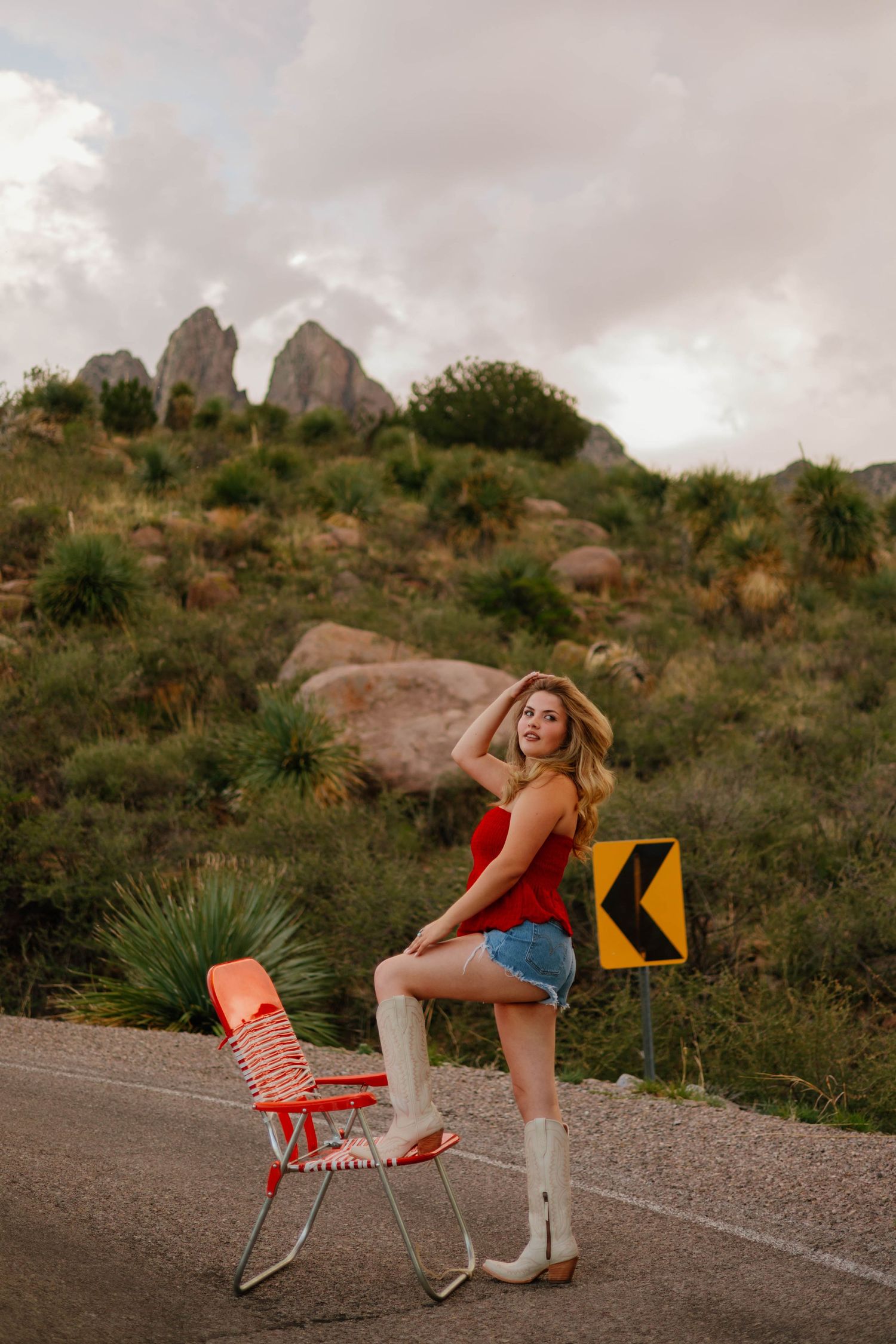Woman in red top, denim shorts, and white cowboy boots posing by a vintage chair on a mountain road.