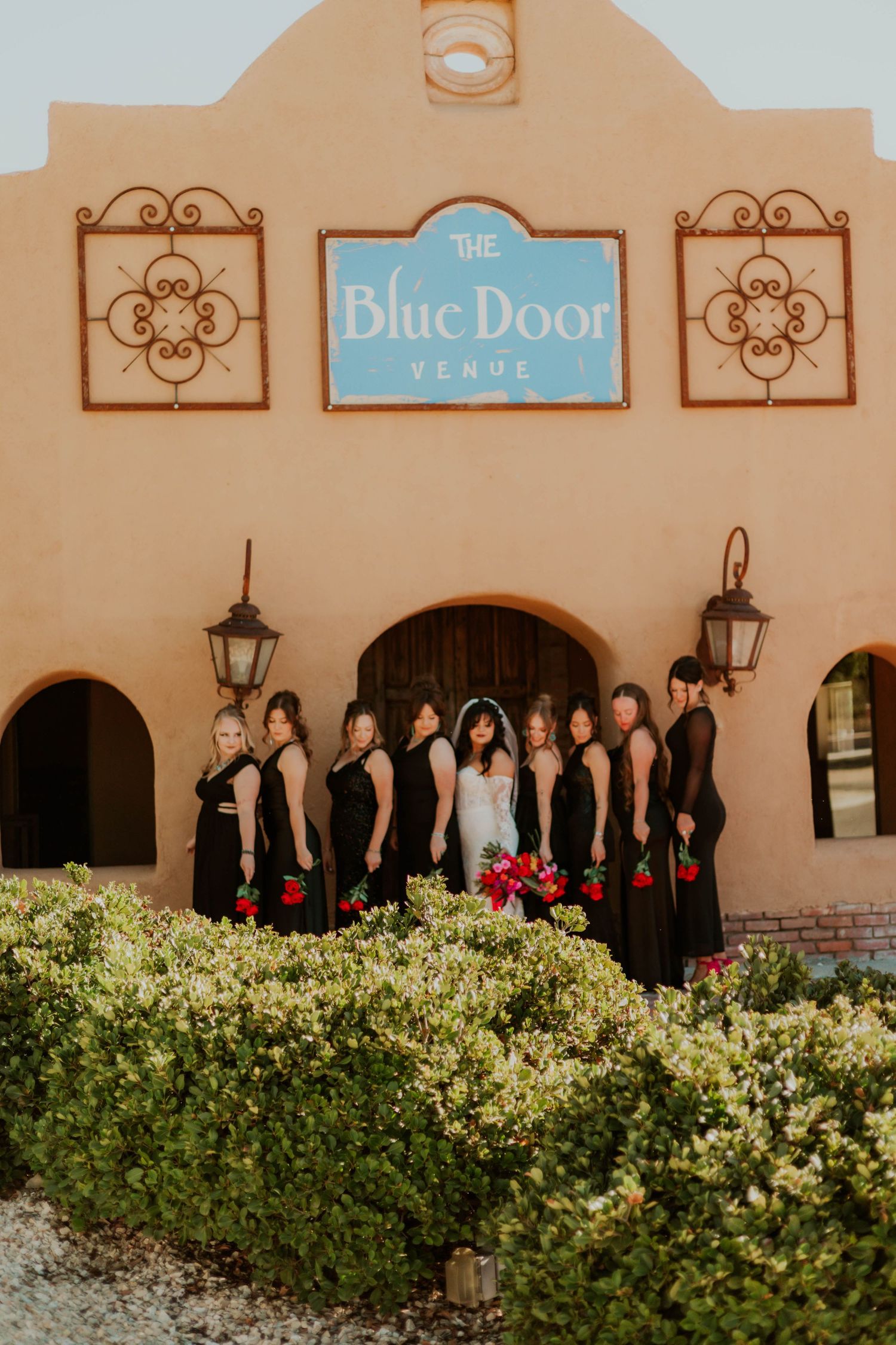 Bride and bridesmaids in black dresses pose in front of The Blue Door Venue, a Spanish-style building with lush greenery.