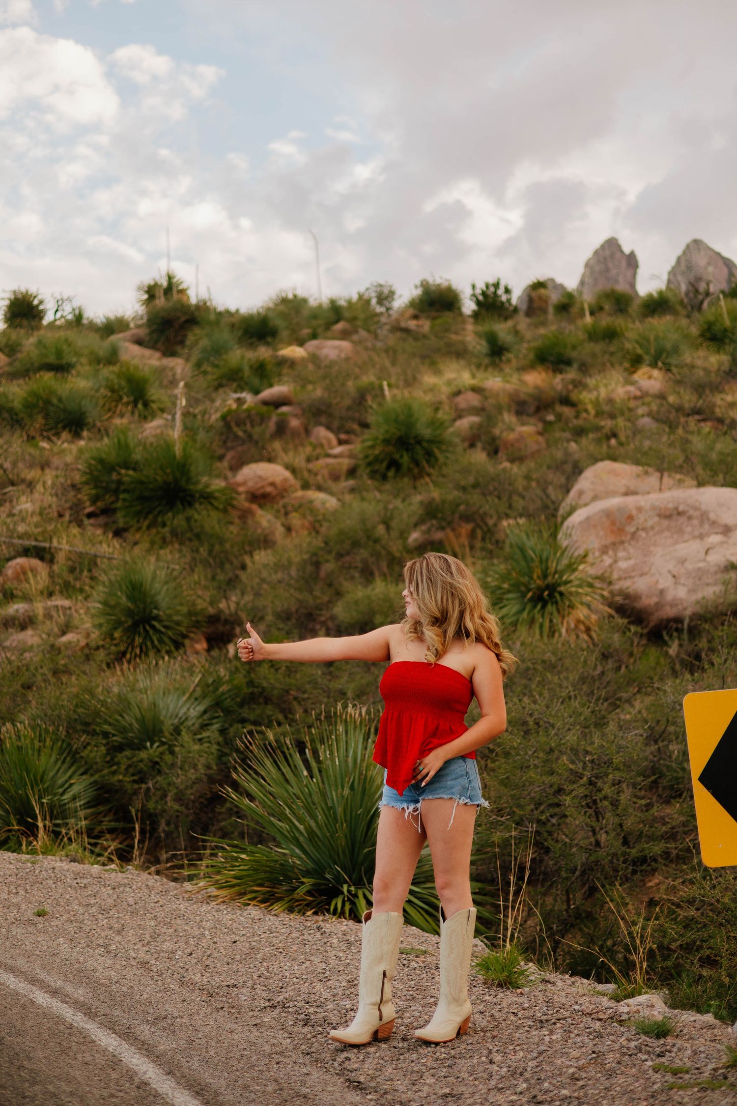 Woman in red top, denim shorts, and white cowboy boots hitchhiking on a desert road with rocky hills.