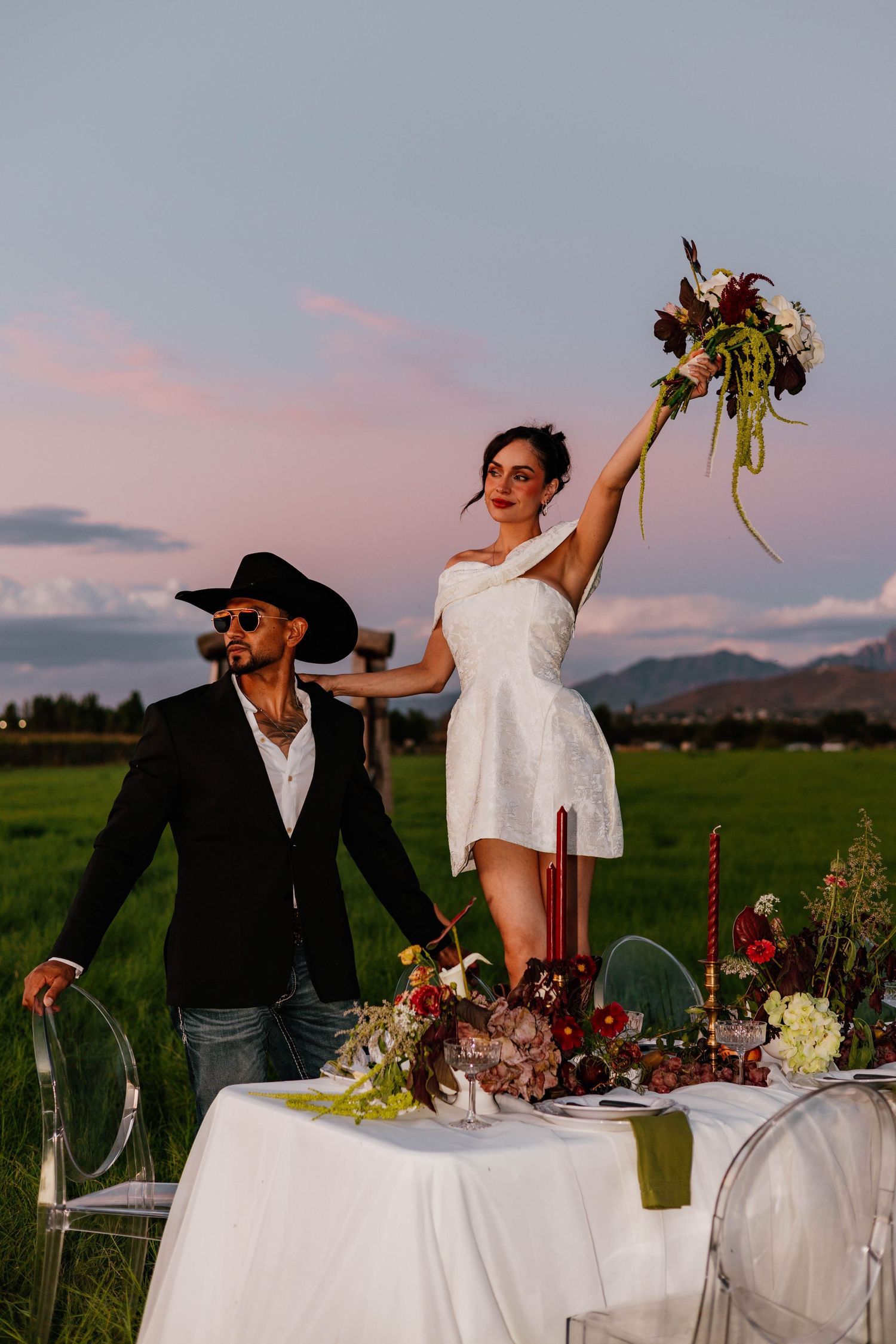 Bride raises bouquet triumphantly beside groom in cowboy hat at outdoor sunset table.
