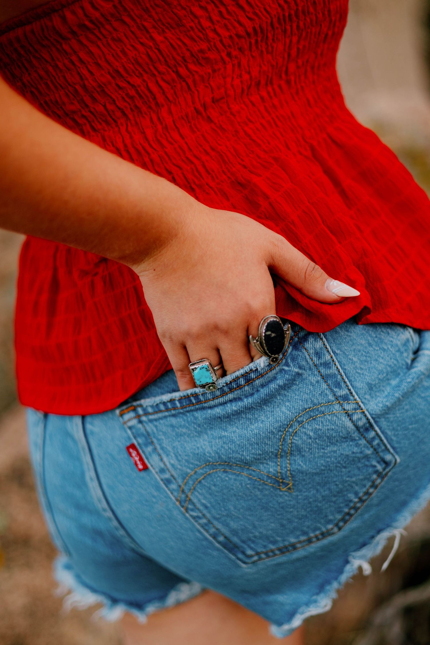 Woman in red smocked top and Levi denim cutoff shorts with turquoise and black rings on her hand.