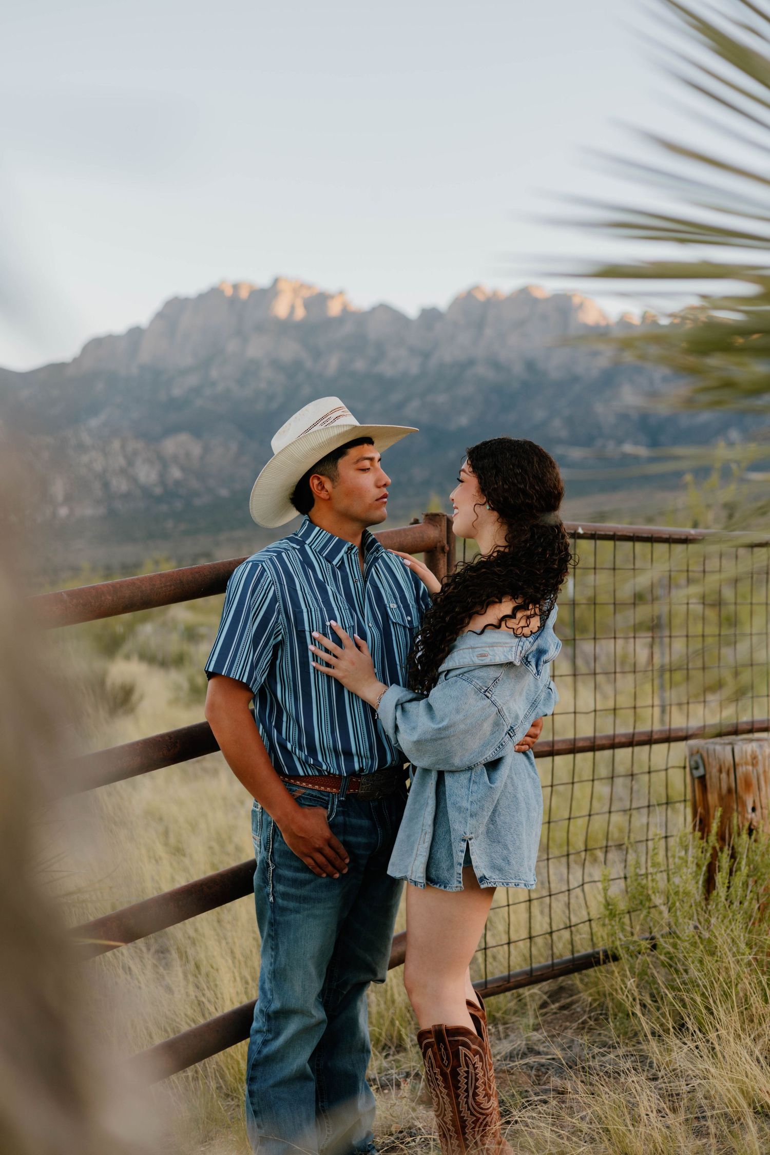 Couple sharing romantic moment by ranch fence with rugged mountain range glowing in background.