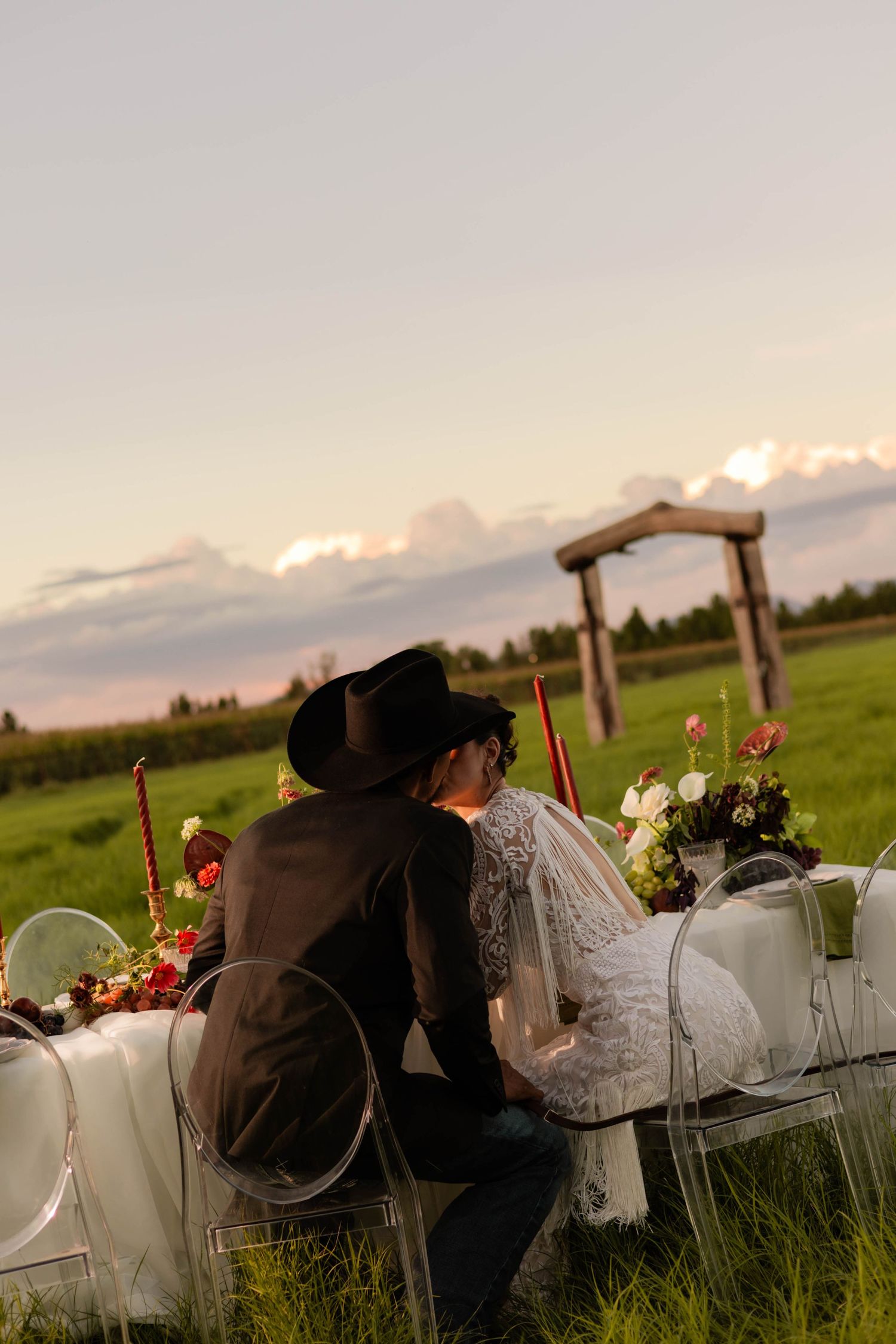 Bride and groom share a romantic outdoor wedding dinner at sunset in a green field with a rustic wooden arch backdrop.