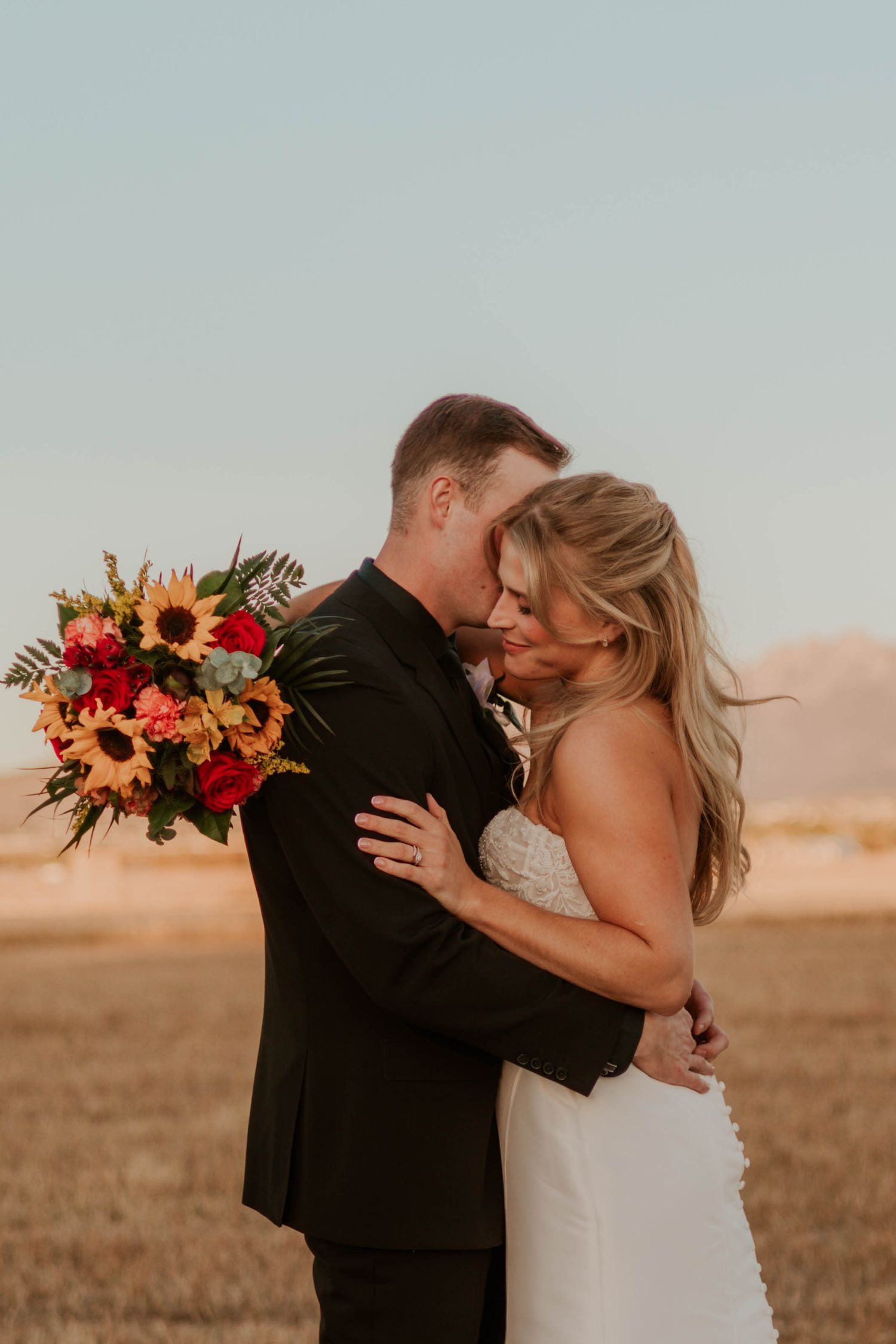 Bride and groom embrace in a golden field, holding a vibrant sunflower and red rose bouquet during sunset.