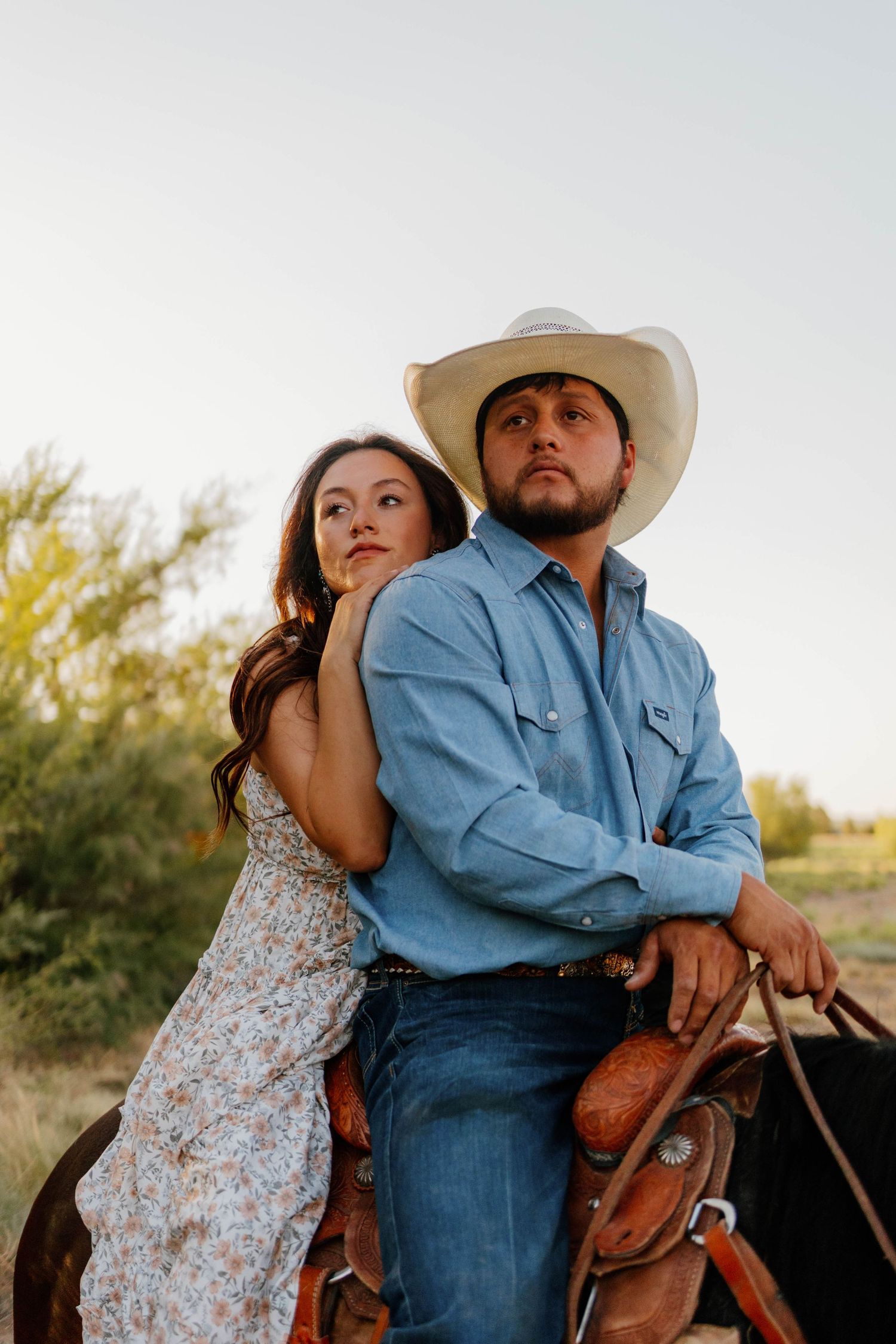 Woman leaning against man in cowboy hat on horseback in golden desert landscape at sunset.