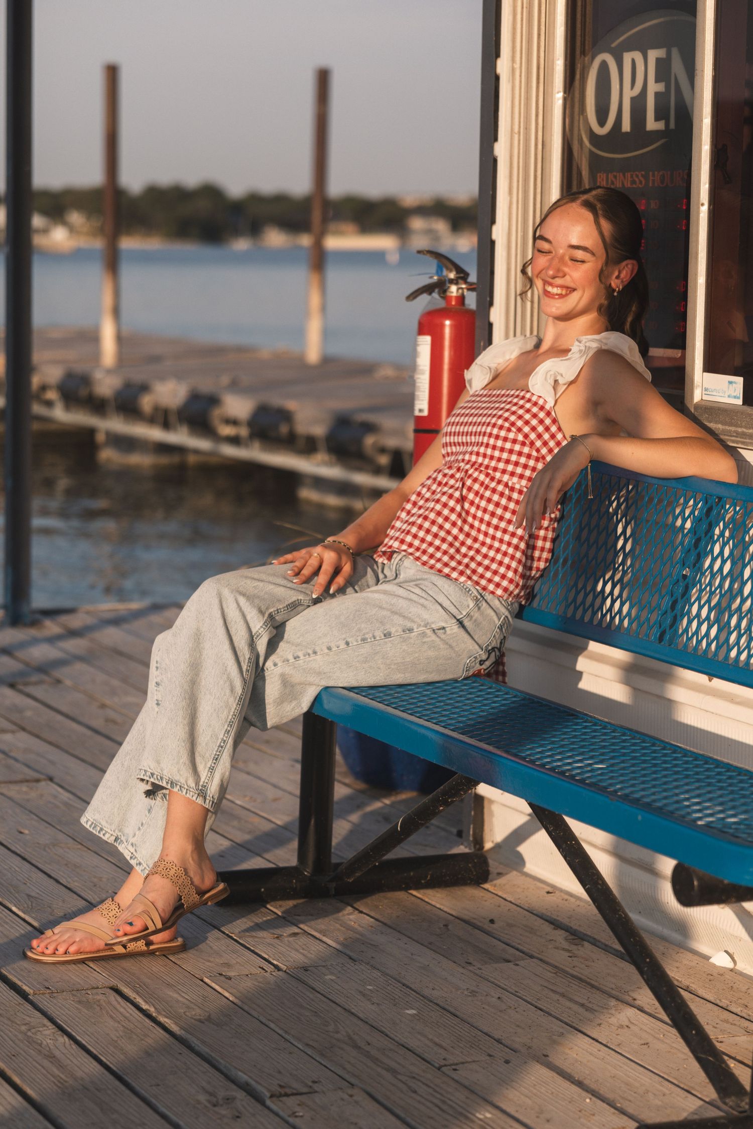 Smiling woman in red gingham top and jeans relaxing on a blue bench on a sunny waterfront dock.