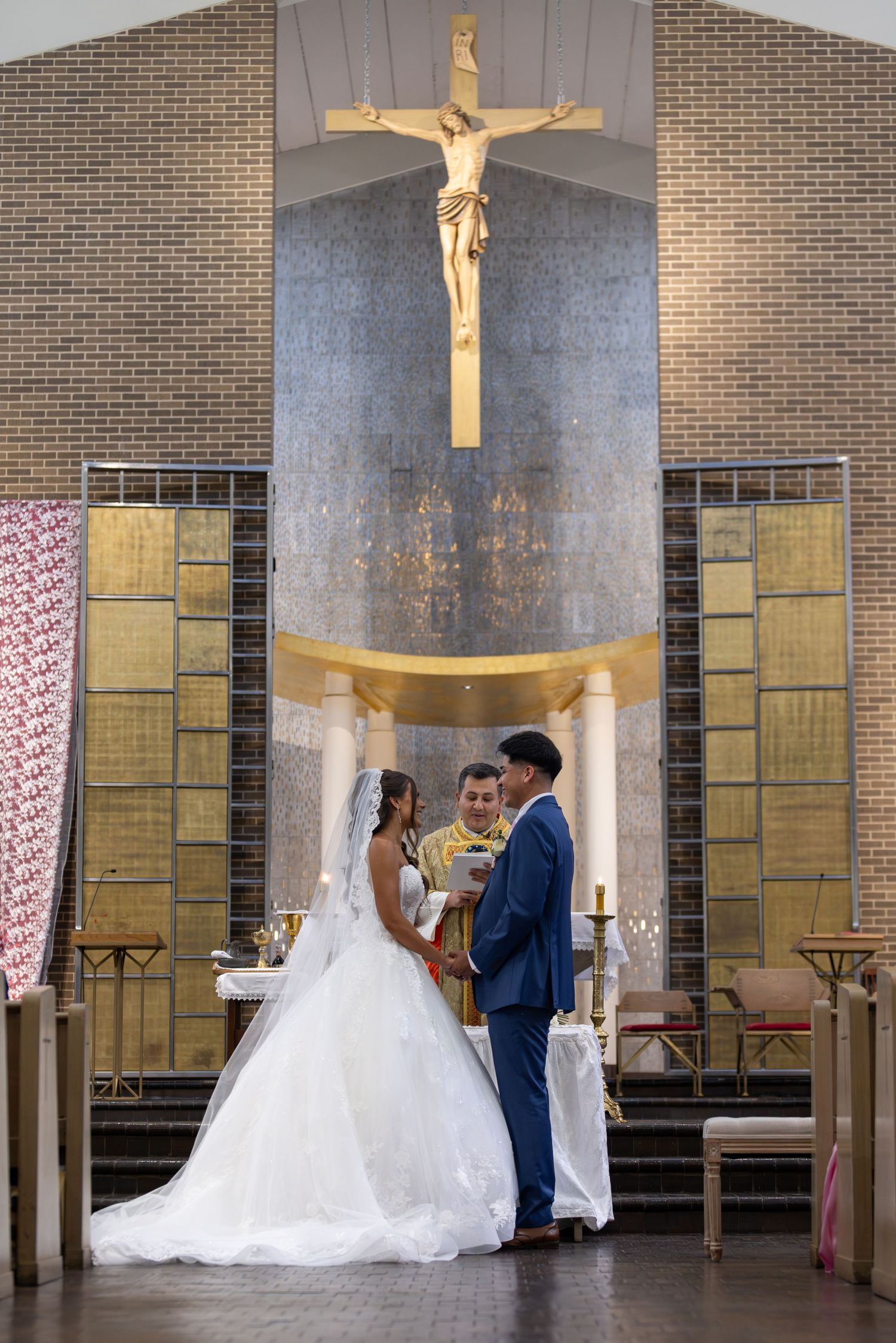Bride and groom exchange vows before a priest at a Catholic church altar with a golden crucifix.