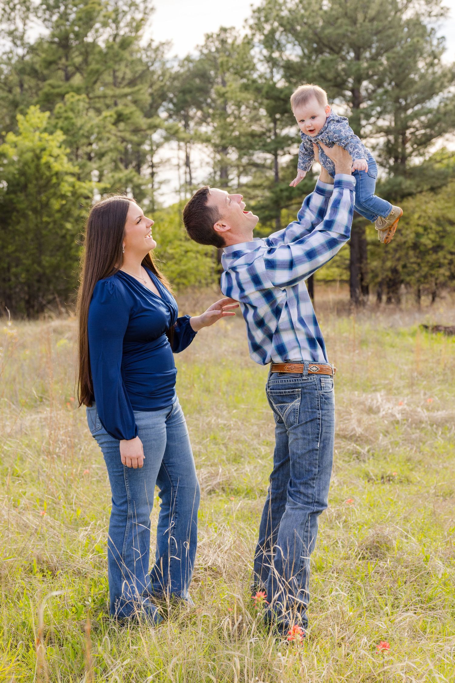 Happy family outdoors, father lifting baby in air while mother watches, standing in grassy field with pine trees.
