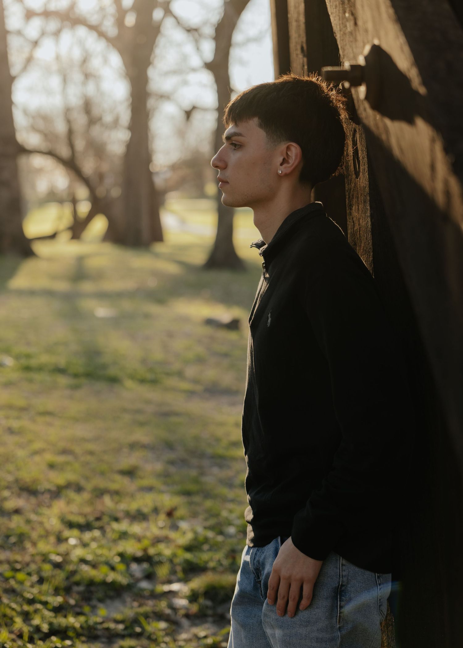Person in a black hoodie and jeans stands near trees in a park during golden hour lighting.