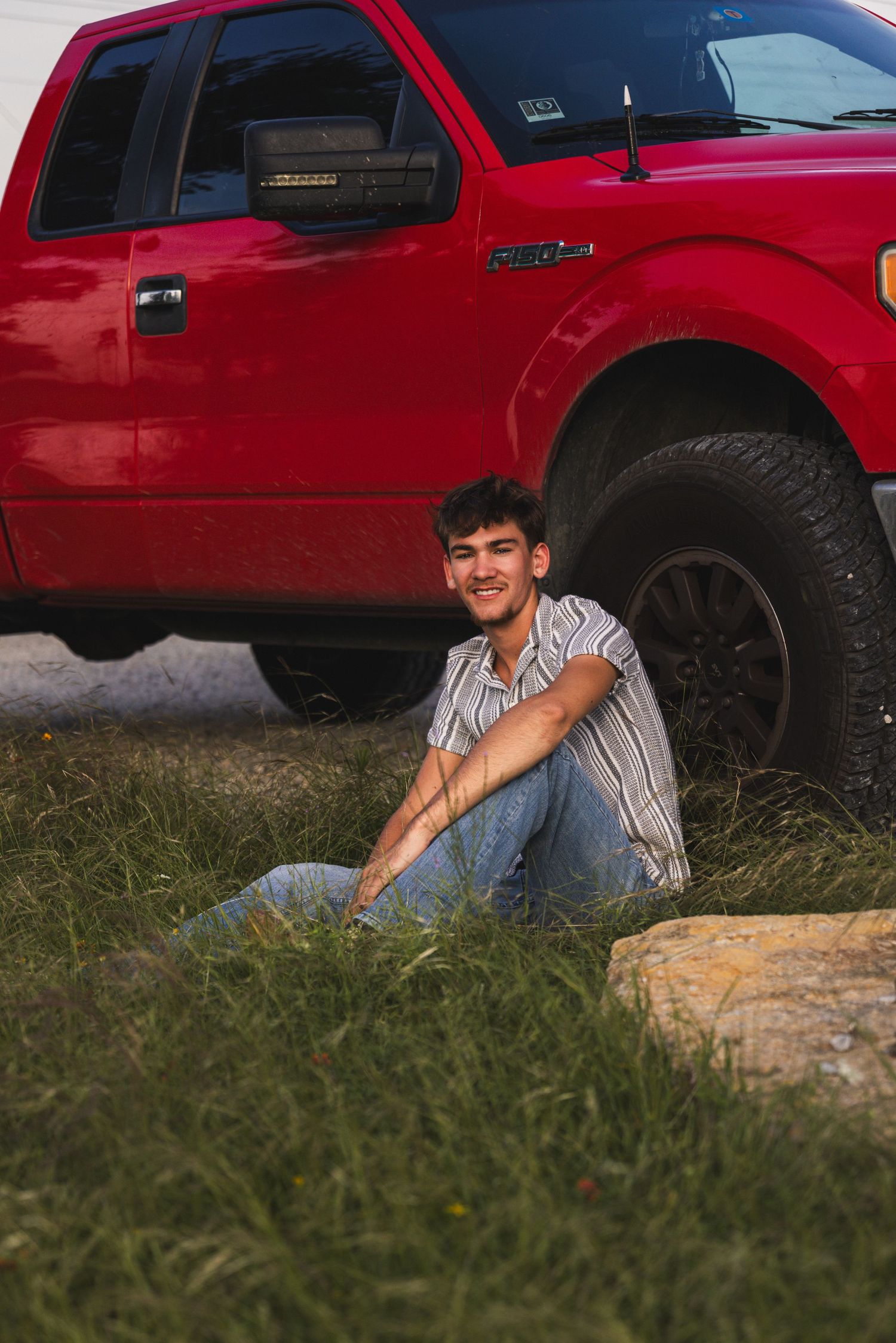 Young man sitting in grass smiling beside a red pickup truck outdoors.