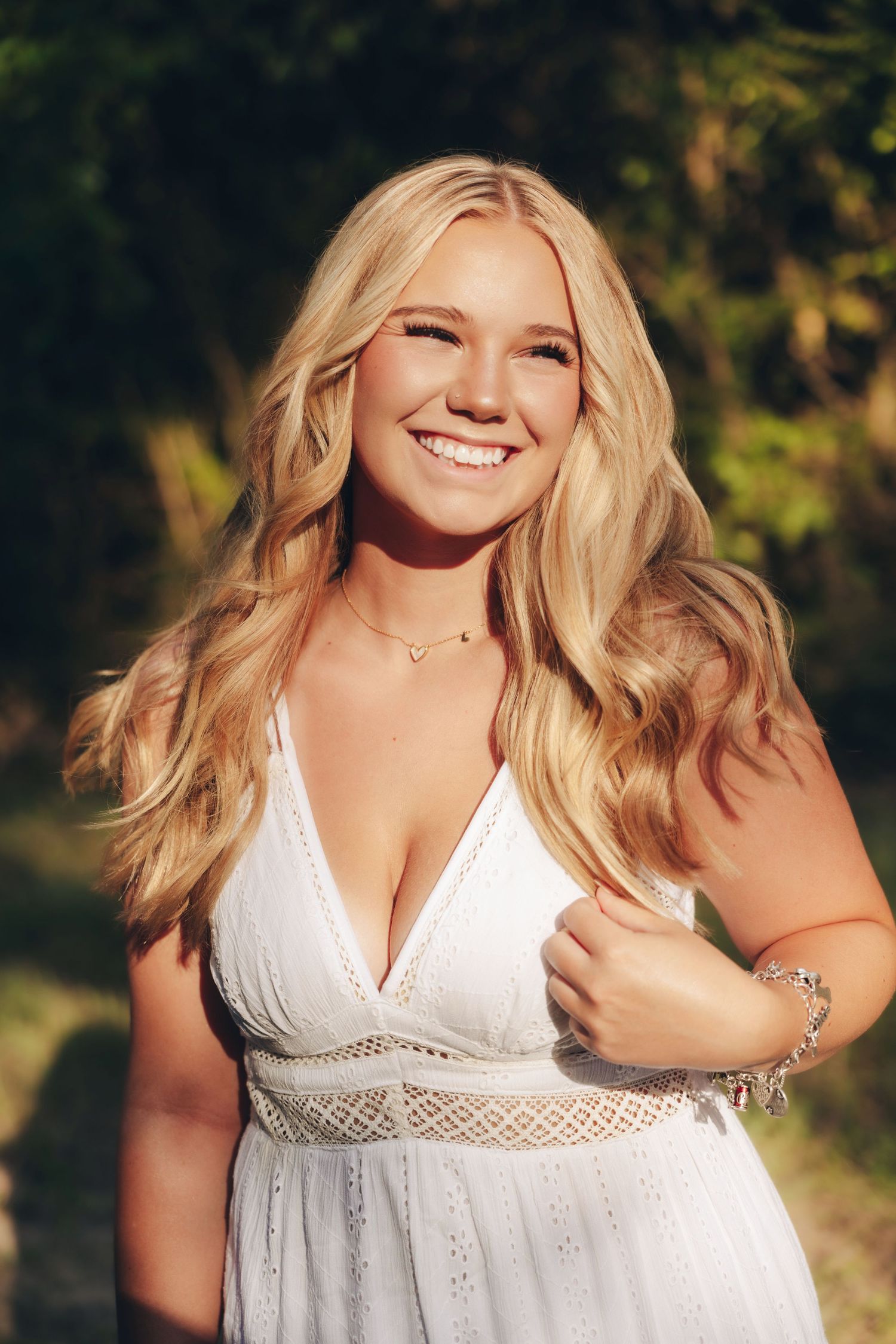 Smiling blonde woman in white sundress with wavy hair, posing outdoors in warm golden sunlight near green trees.