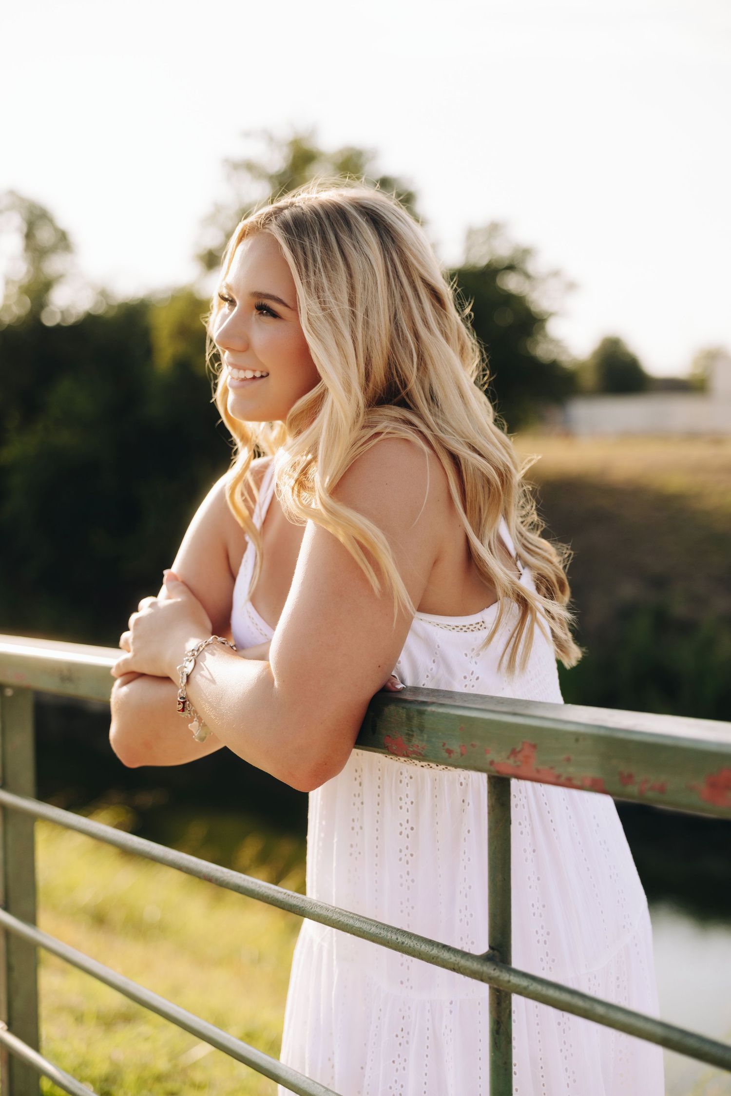 Smiling blonde woman in white dress leaning on a green fence outdoors in golden hour sunlight.