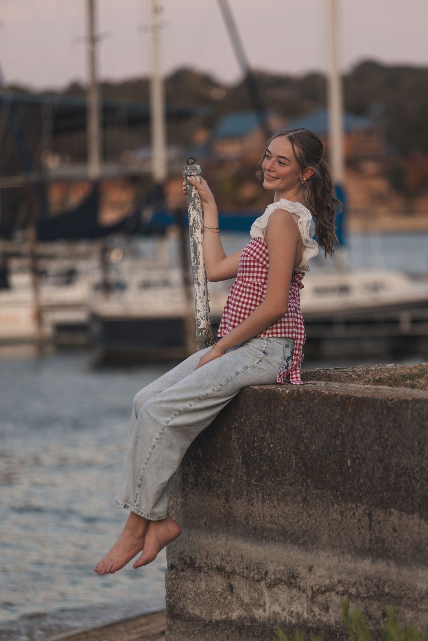 Young woman in checkered top and jeans sits barefoot on a harbor wall, smiling while holding fabric near sailboats.