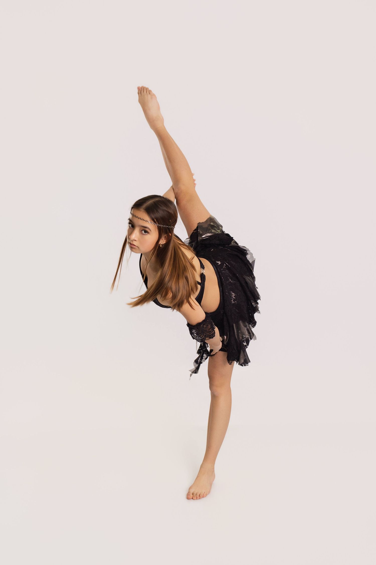 Young girl in black dance costume performs a standing split pose against a white background.