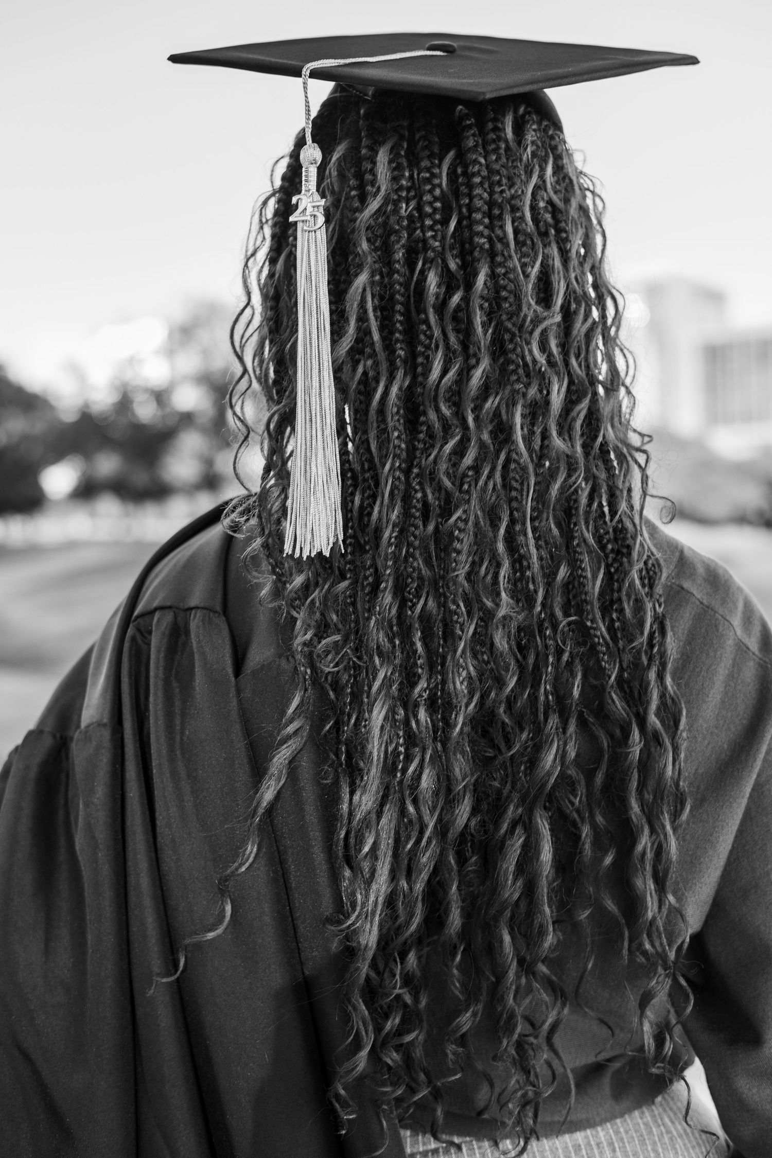 Back view of graduate wearing black cap and gown with long curly hair and white tassel at commencement ceremony.