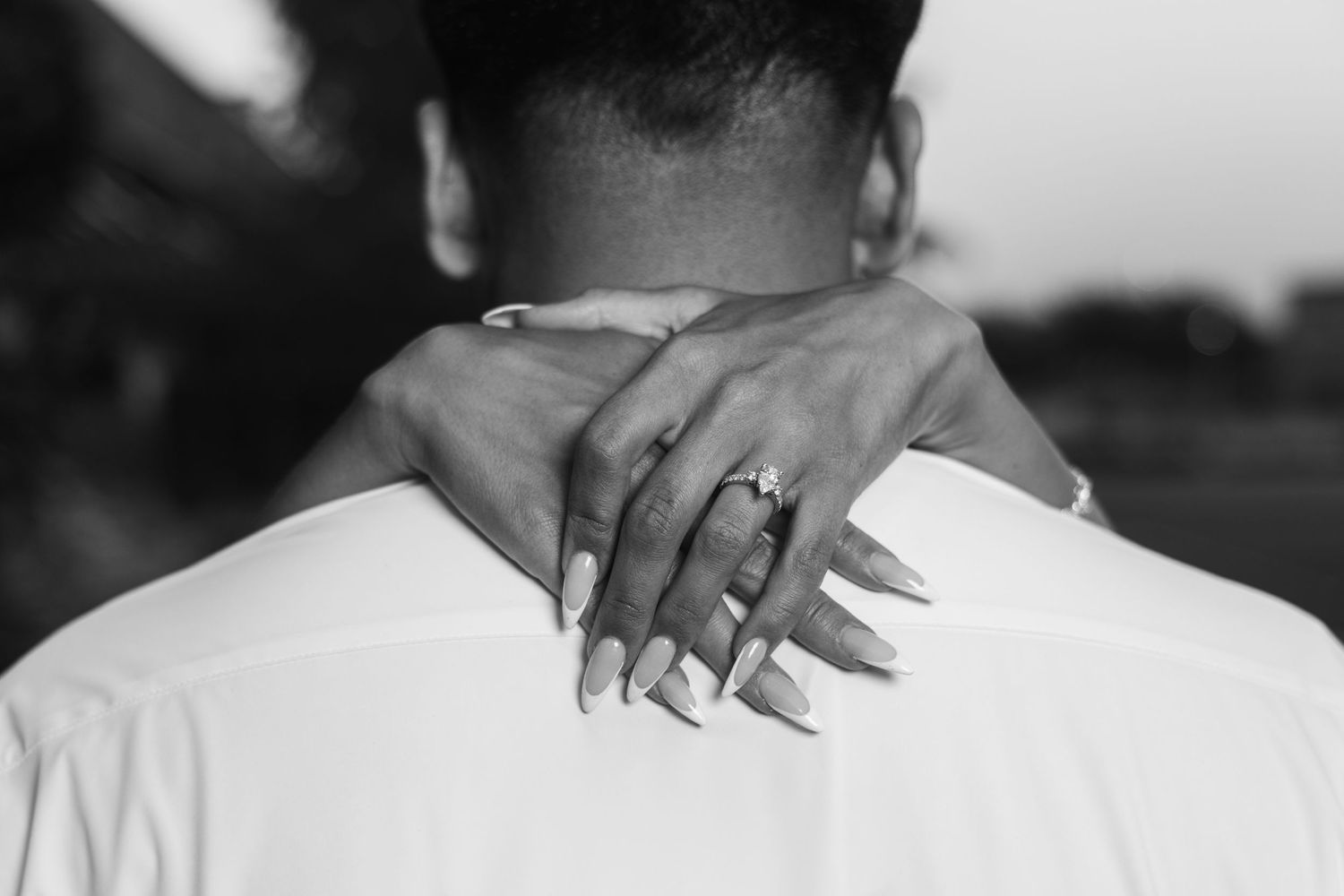 Black and white photo of a woman's hand with engagement ring resting on a man's shoulder.