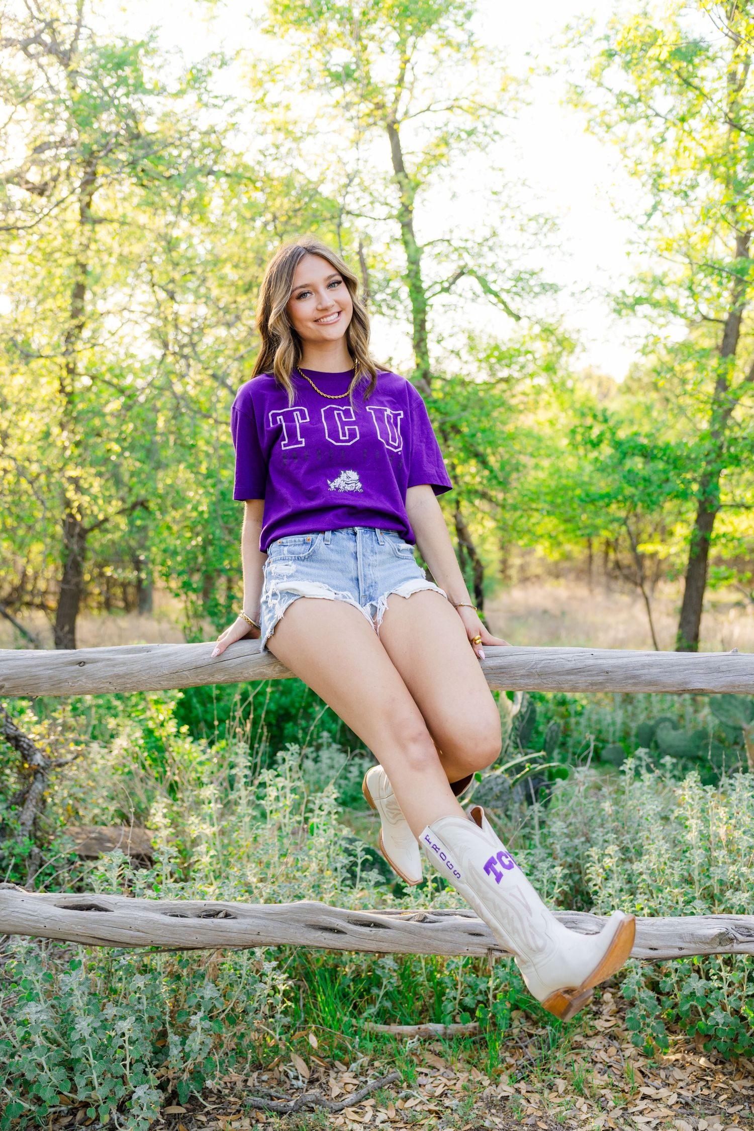 Young woman in TCU purple t-shirt, denim shorts, and cowboy boots sitting on a wooden fence outdoors.