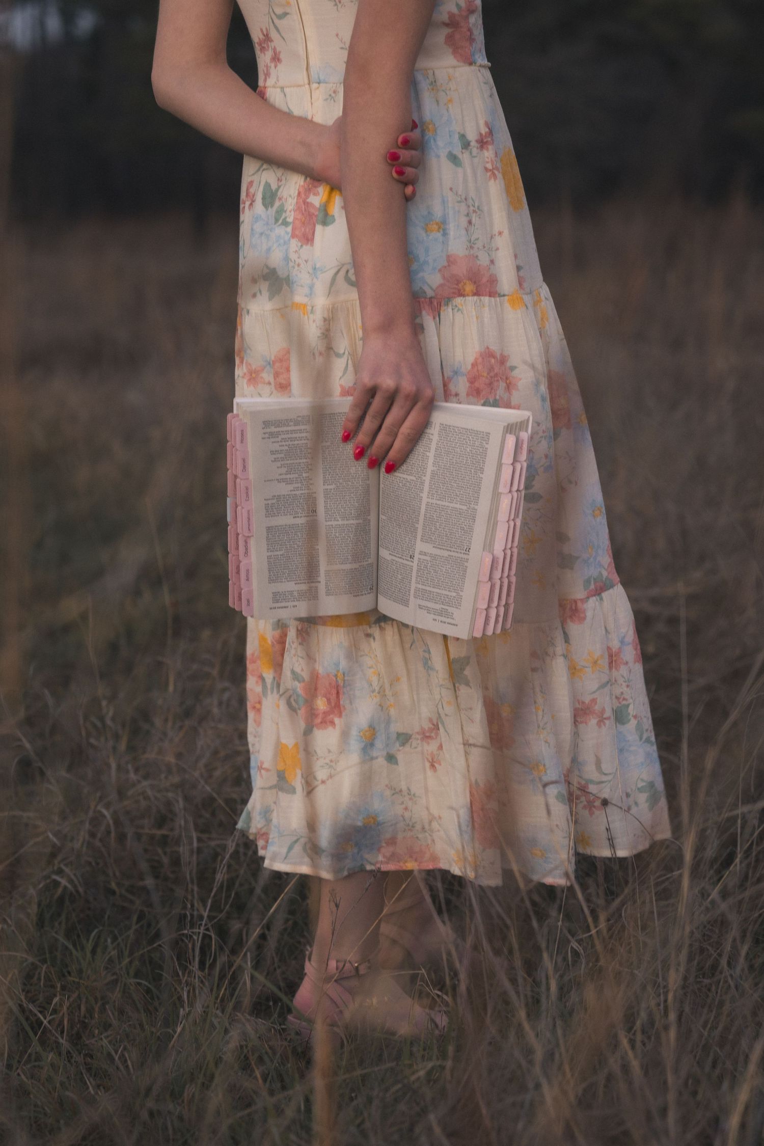 A person in a floral dress stands in tall grass at dusk, holding a book with pages spread open.