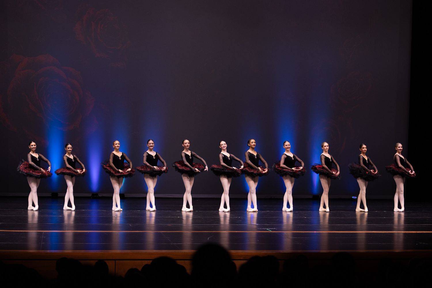 A line of ballet dancers in black costumes perform in unison on a blue-lit stage during a recital.