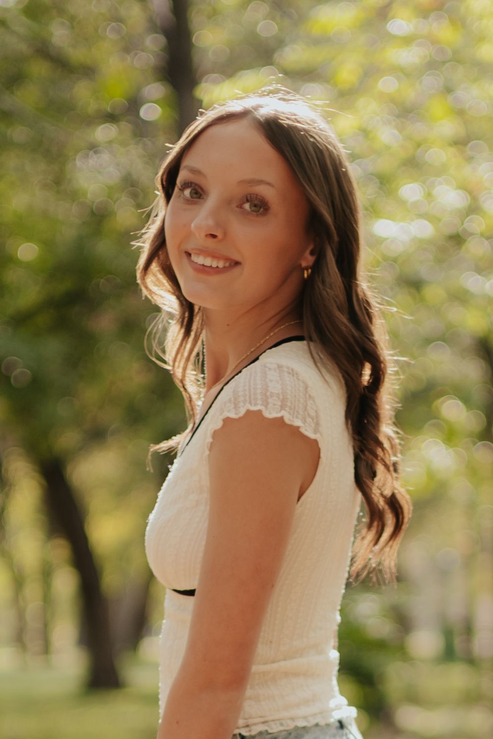 A person in a white dress spins playfully in a sun-dappled park setting with bokeh lighting effects in the background.