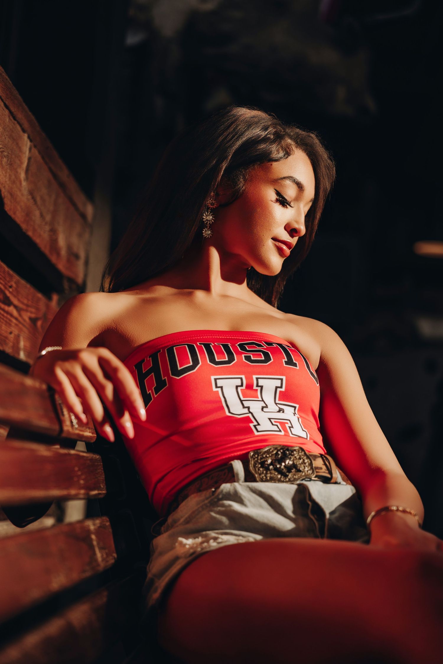 Young woman in red Houston UH tube top and denim shorts seated on wooden bench in dramatic lighting.