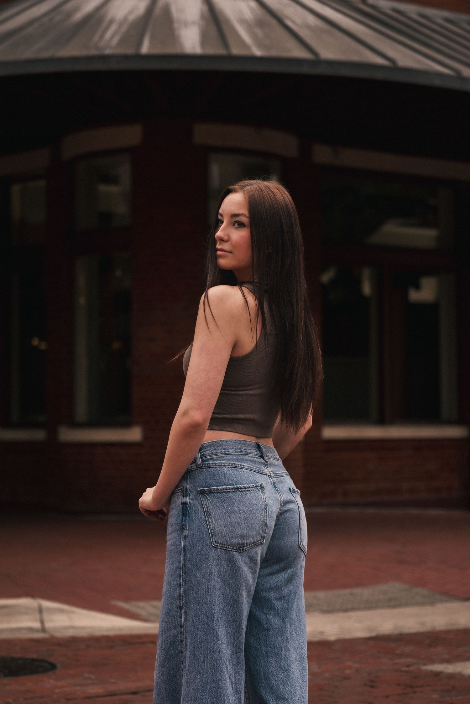 Young woman with long dark hair poses outdoors in crop top and wide-leg jeans near brick building.